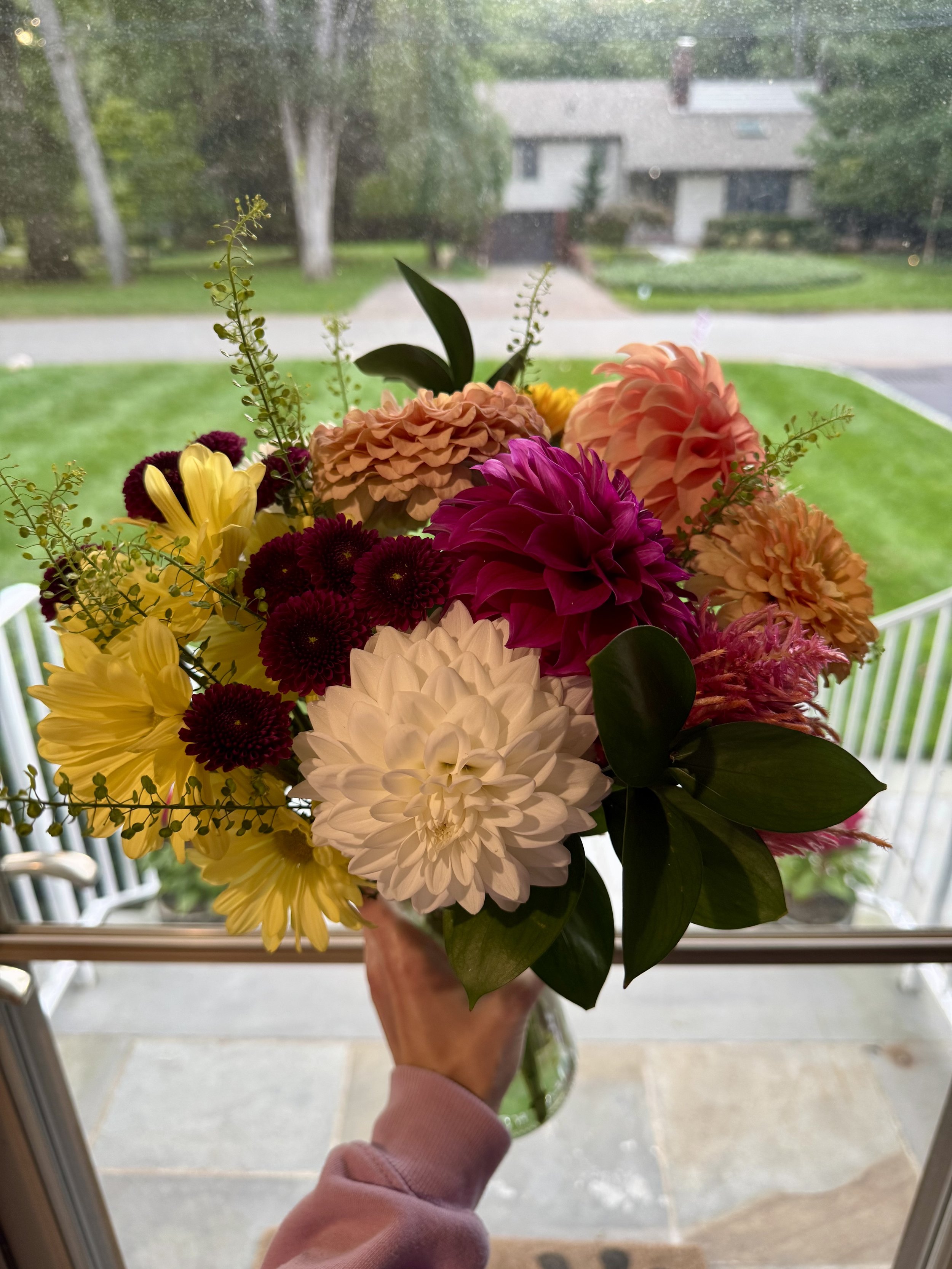 Person holding a bouquet of multicolored flowers in front of a window showing a suburban neighborhood with trees and houses.