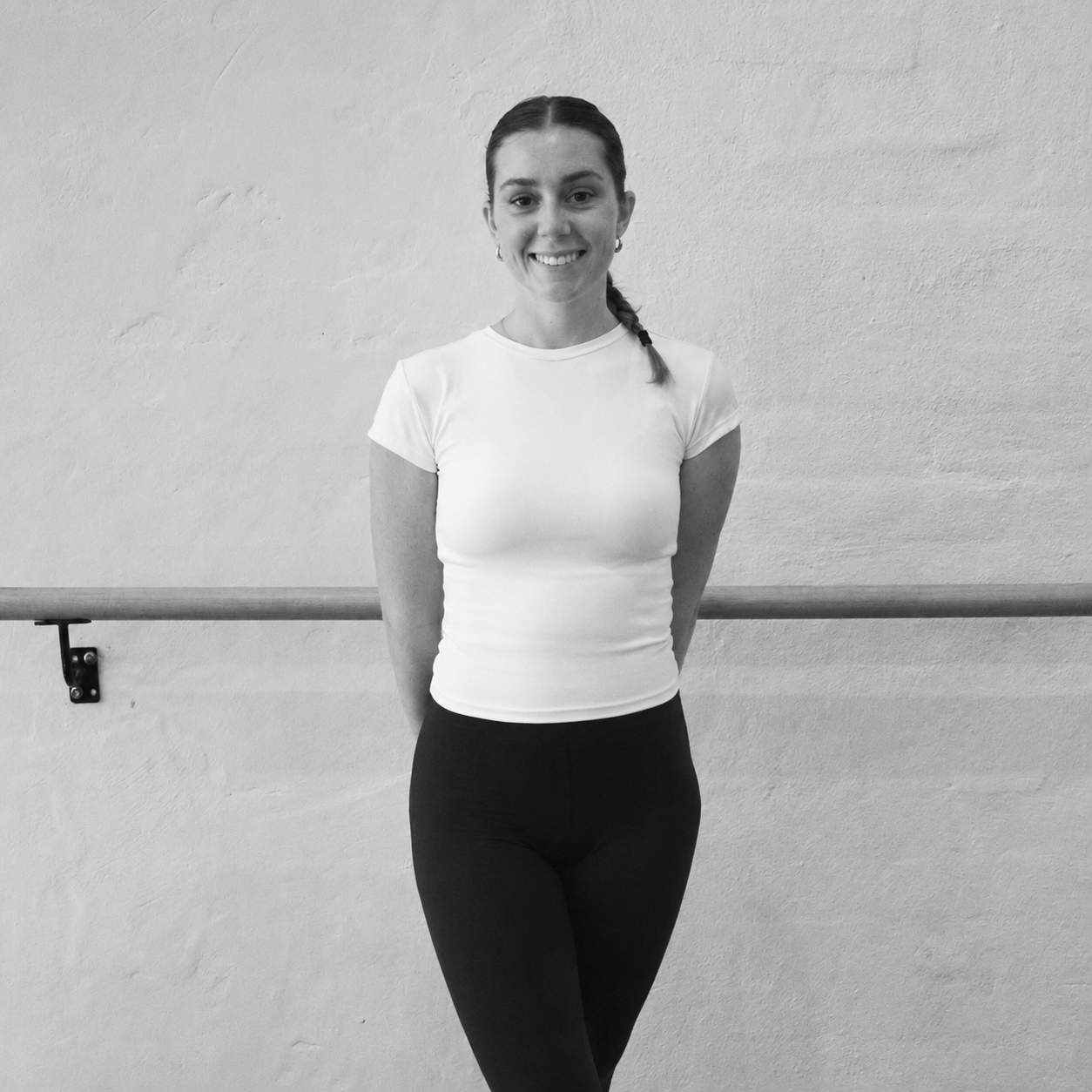A smiling woman with long hair tied back, wearing a white t-shirt and black leggings, standing in front of a ballet barre on a textured wall.