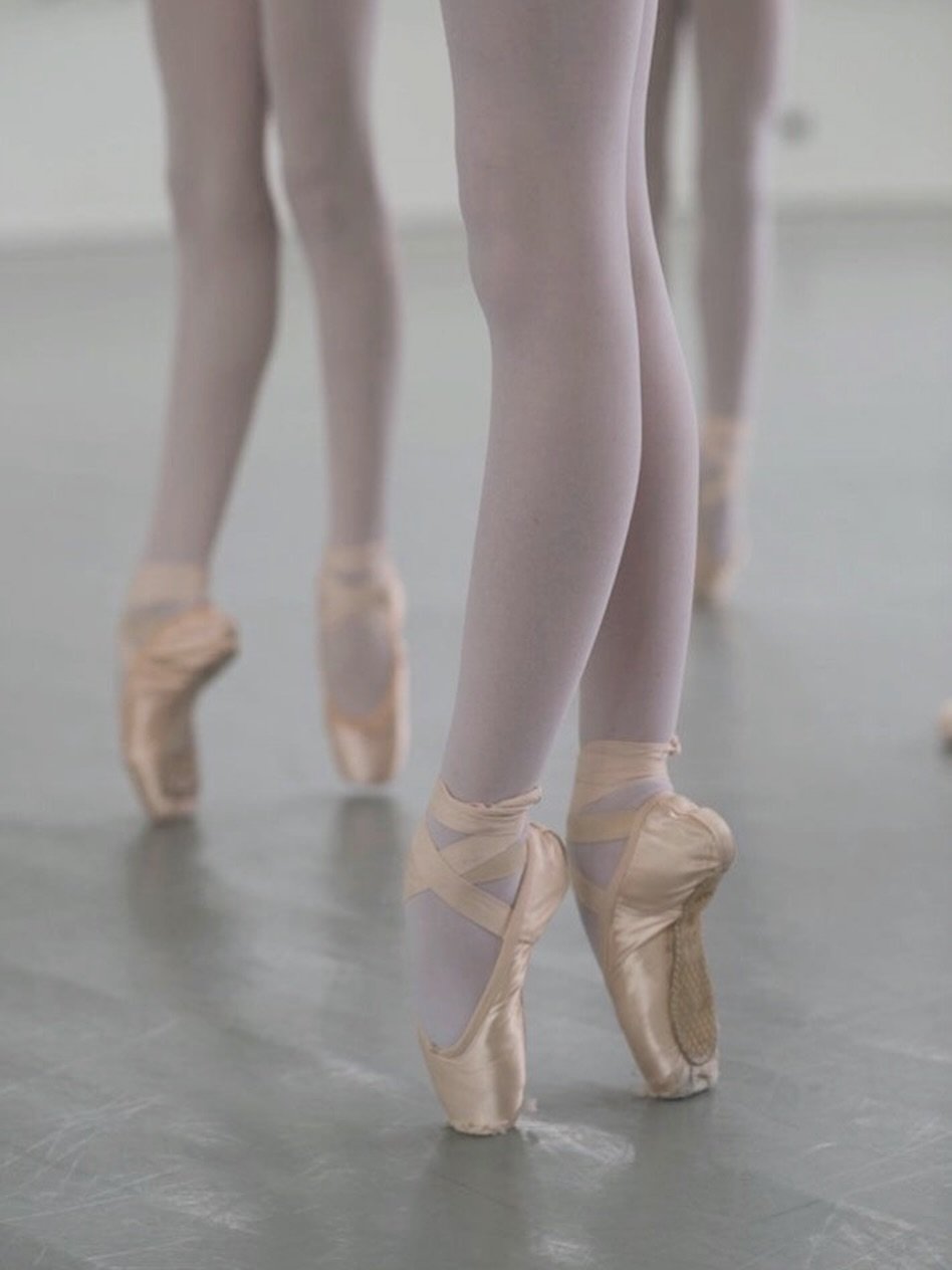 Close-up of ballet dancers' feet in pointe shoes and white tights, positioned in a ballet studio.