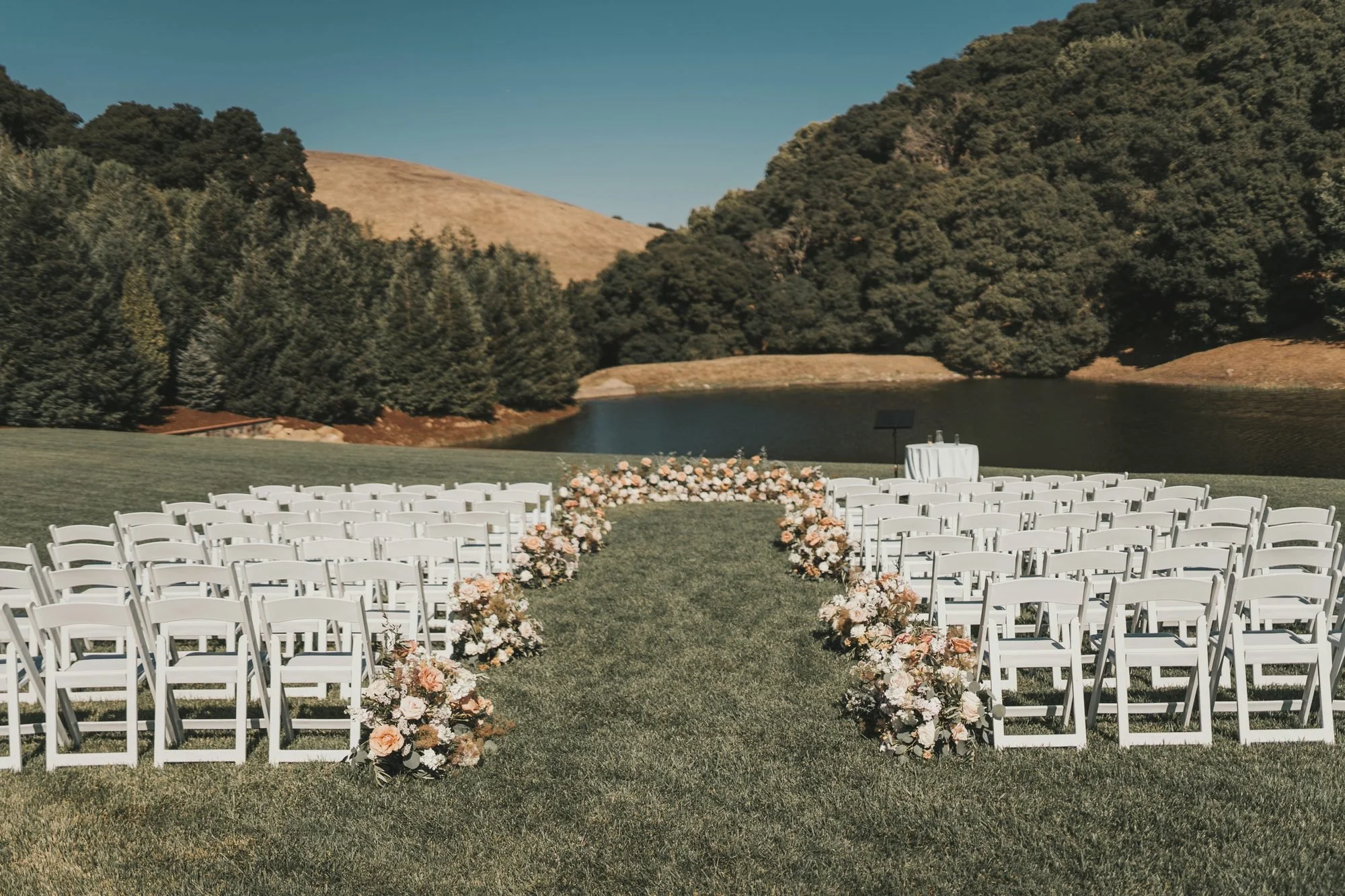 White garden chairs lining a peach and ivory floral aisle beside a lake and golden hills