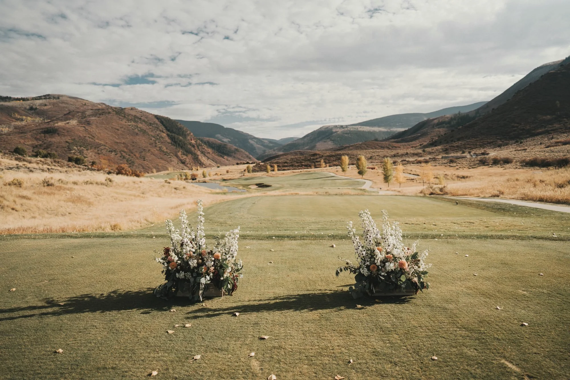 Two white delphinium floral arrangements framing a golf-course ceremony site with autumn mountain views