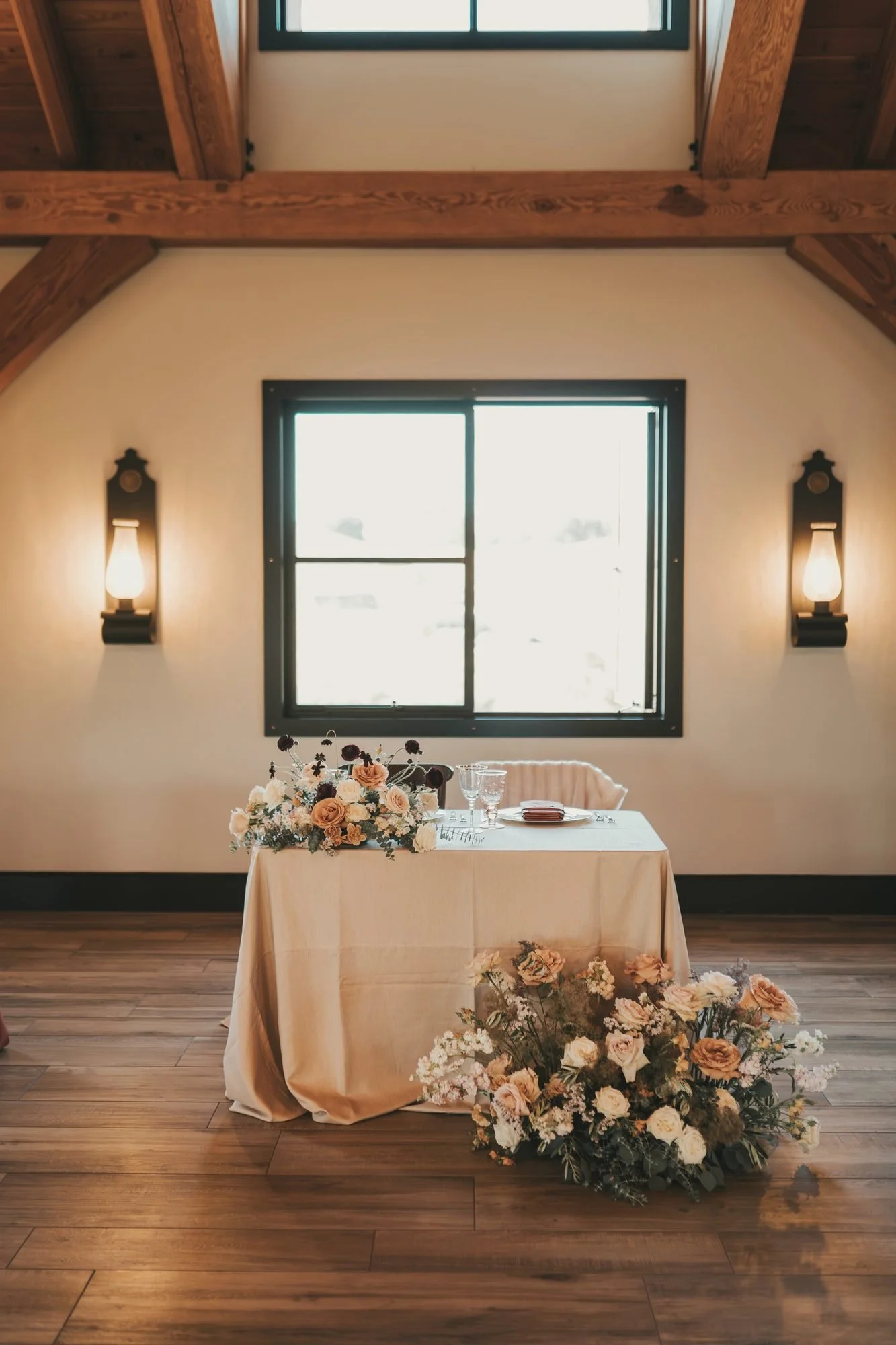 Sweetheart table draped in champagne linen with peach florals beneath a window and glowing sconces