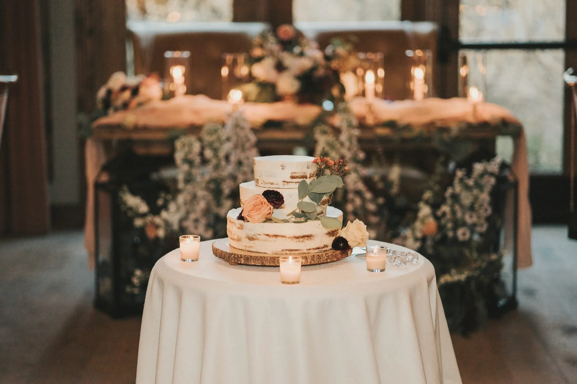 Three-tier naked cake with roses and eucalyptus on a candlelit table before a delphinium sweetheart table