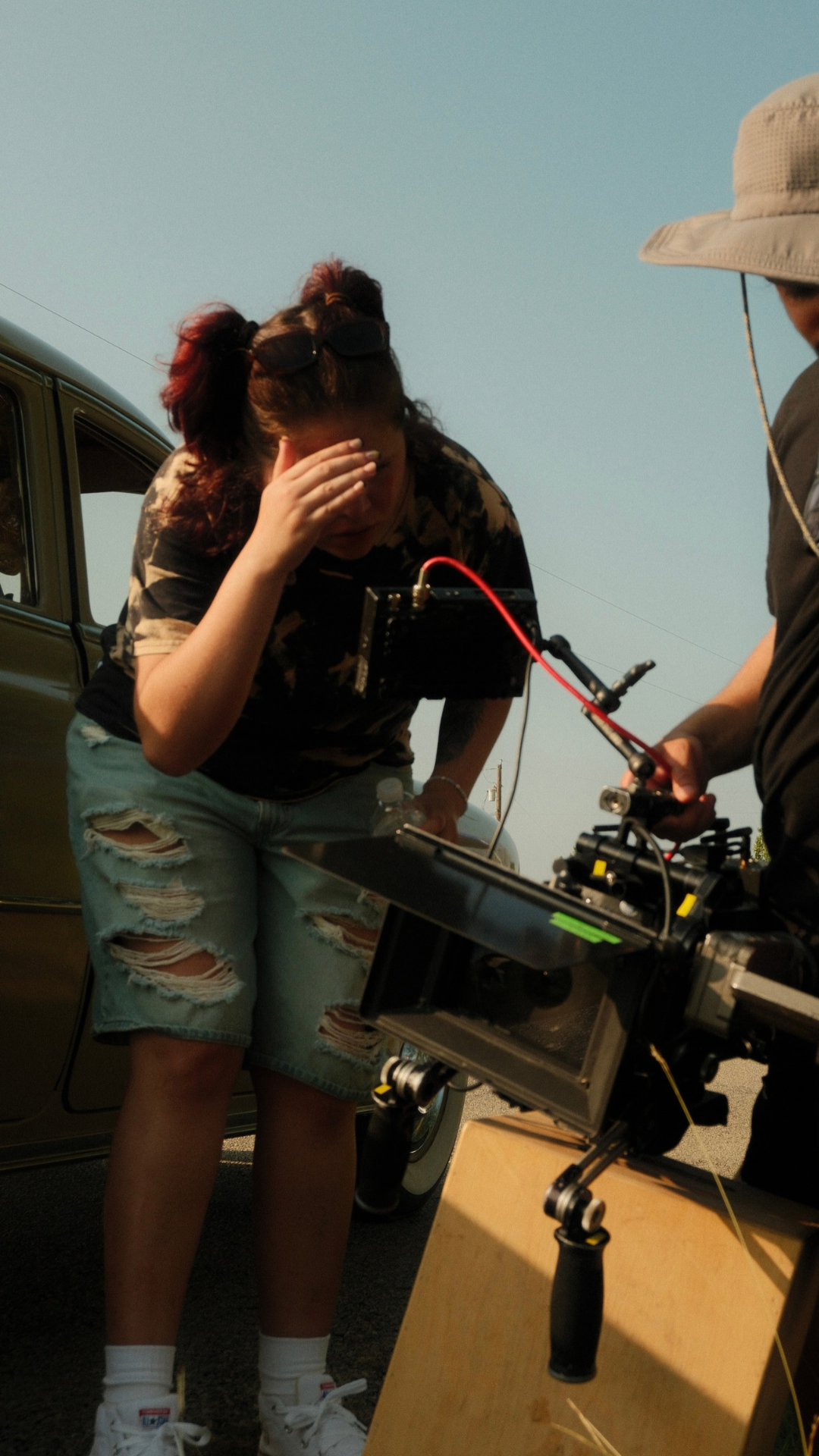 Film crew member shielding her eyes beside a vintage car while a camera operator adjusts a professional video camera outdoors
