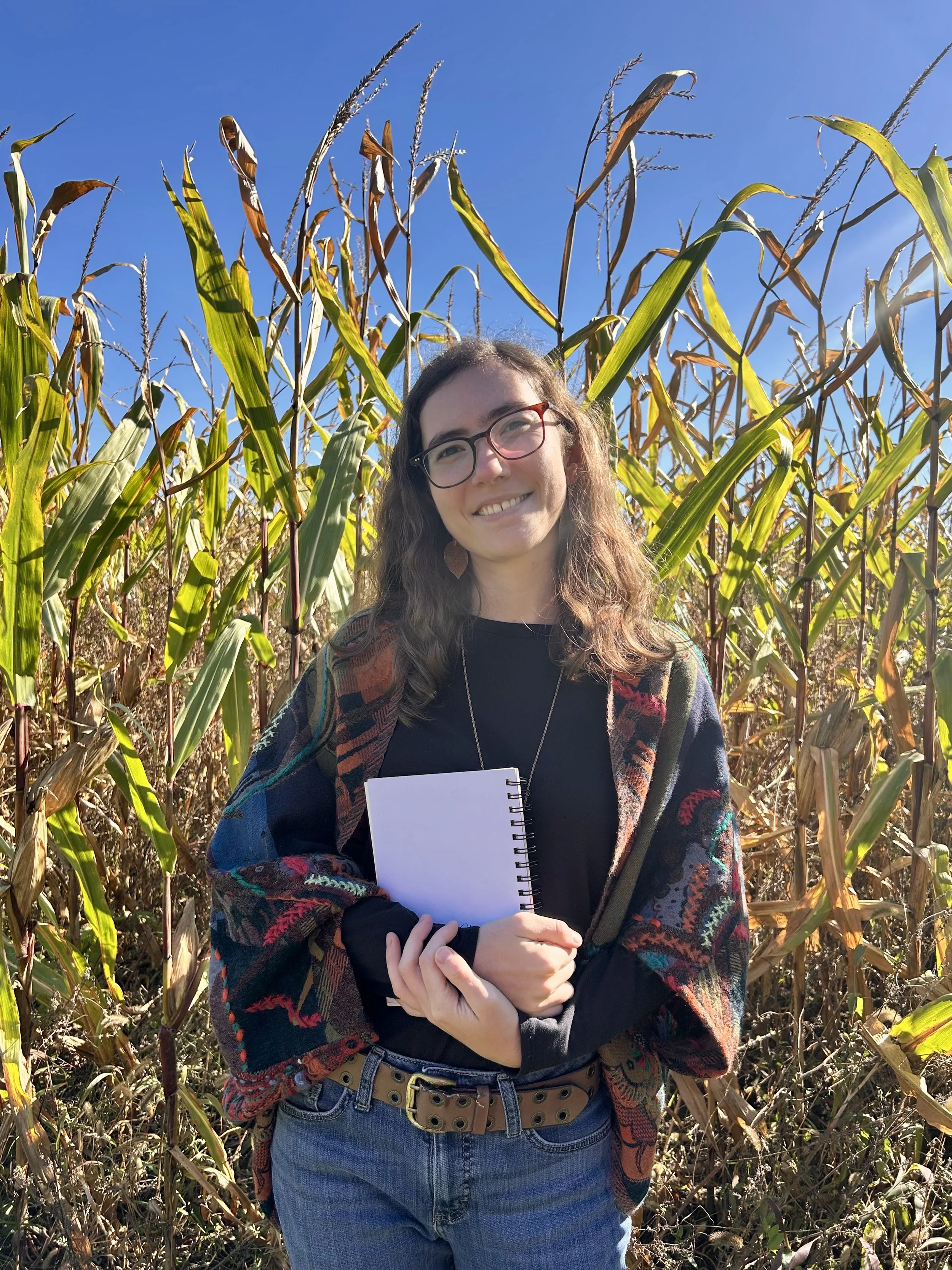 A young woman with glasses and long wavy hair smiling while standing in a cornfield on a sunny day, holding a spiral notebook.