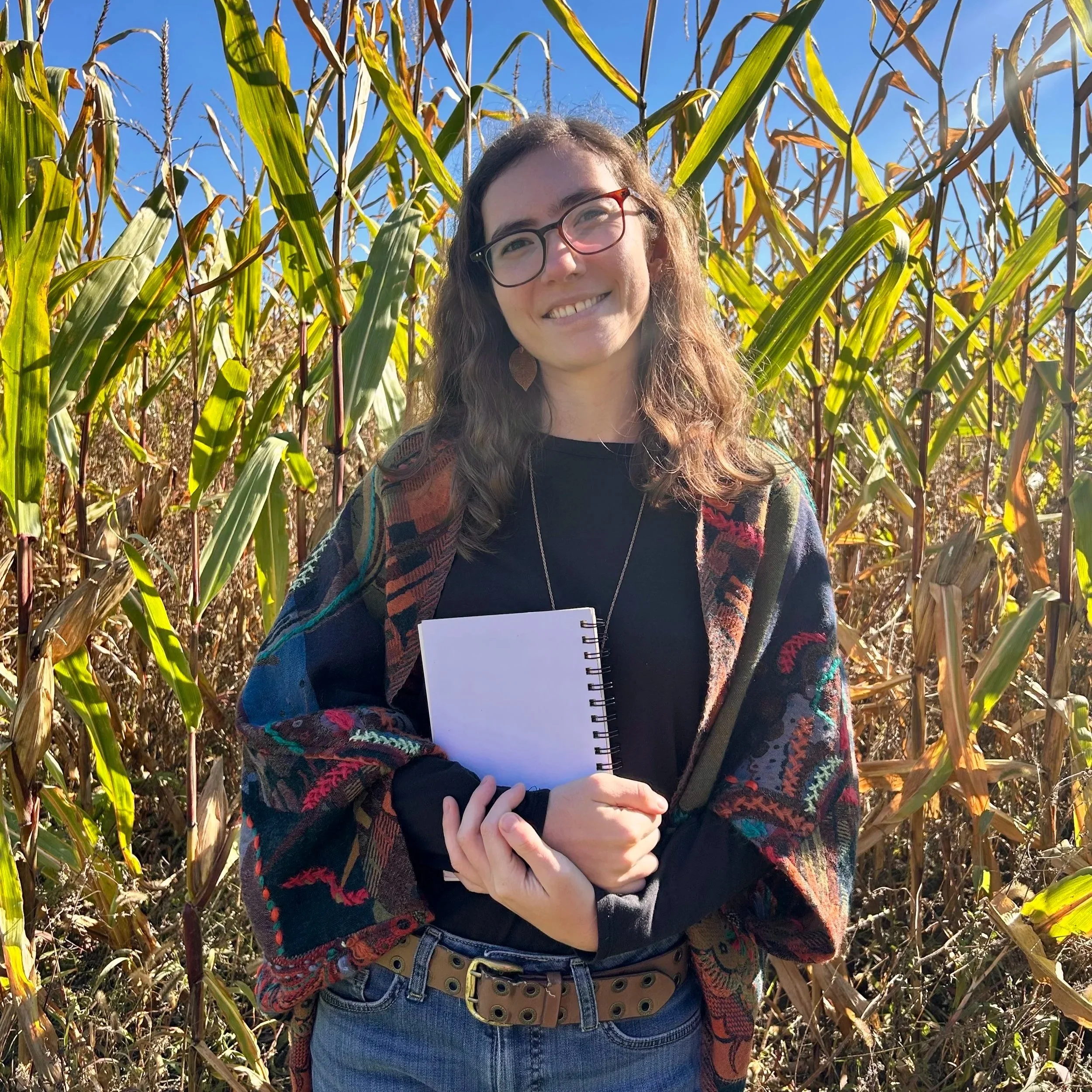 A woman smiling and holding a notebook while standing in a cornfield during daytime with clear blue sky.