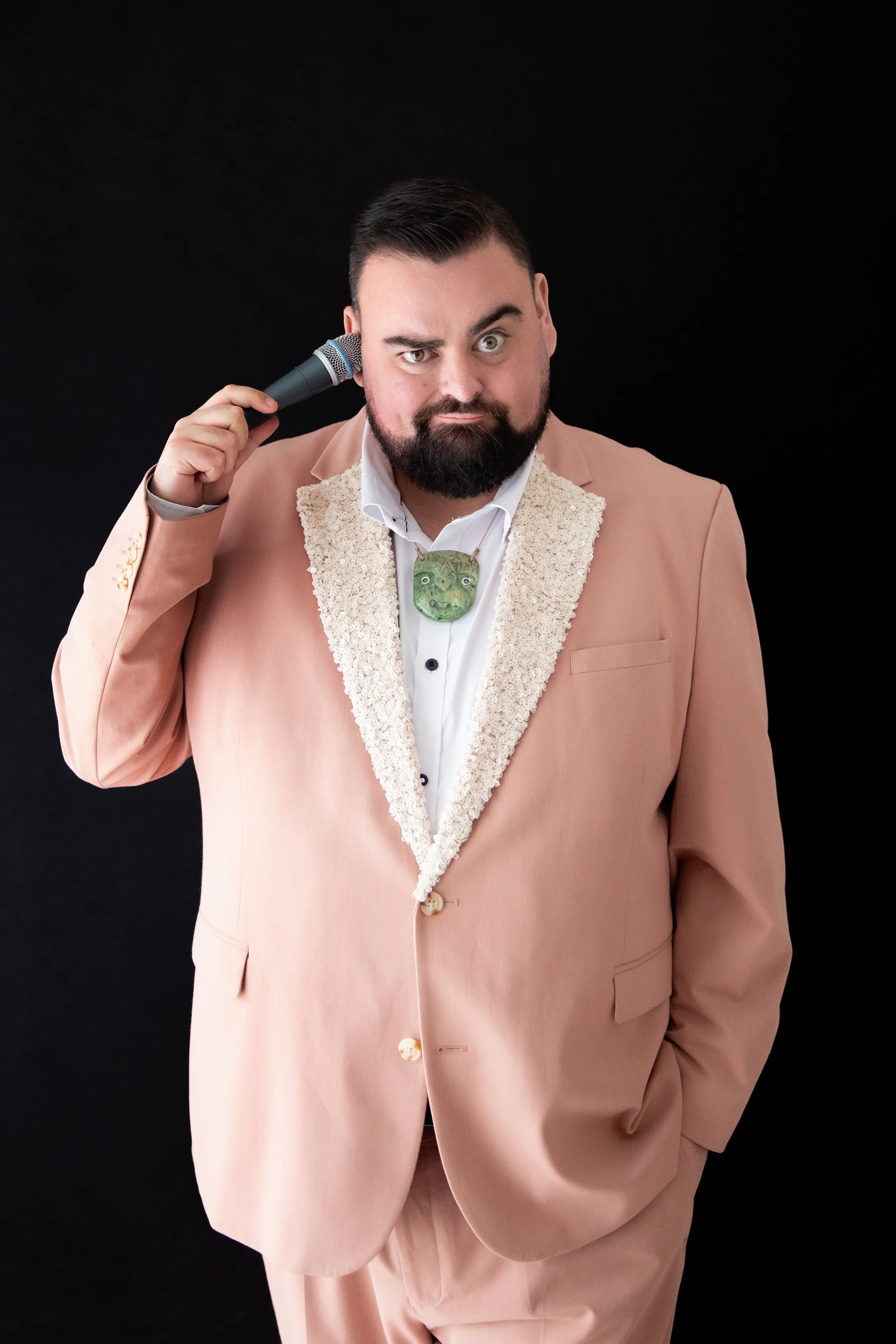 A man with a beard and short dark hair wearing a pink suit with a textured collar, holding a microphone to his head, against a black background.