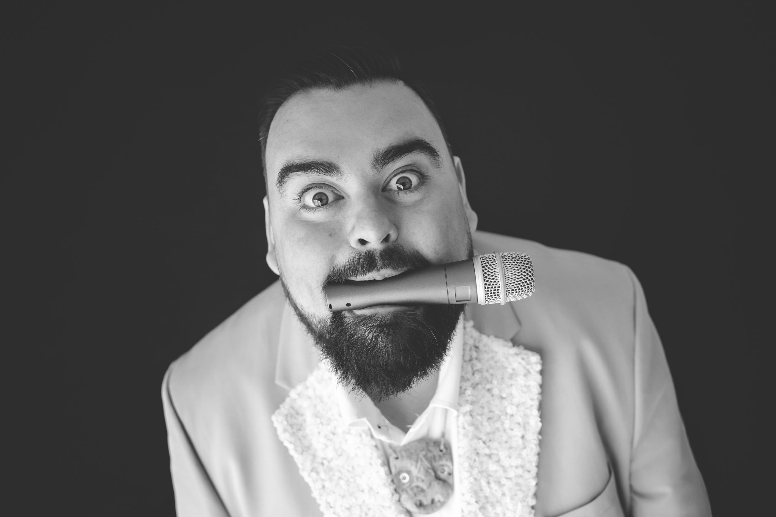 Black and white photo of a man with a beard and mustache, looking directly at the camera, with wide eyes and raised eyebrows, holding a microphone close to his mouth.