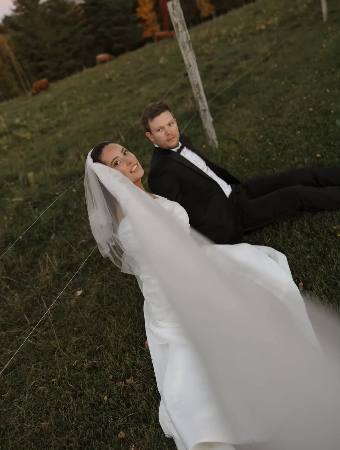 Bride in a white wedding gown and groom in a black tuxedo reclining on grass outdoors