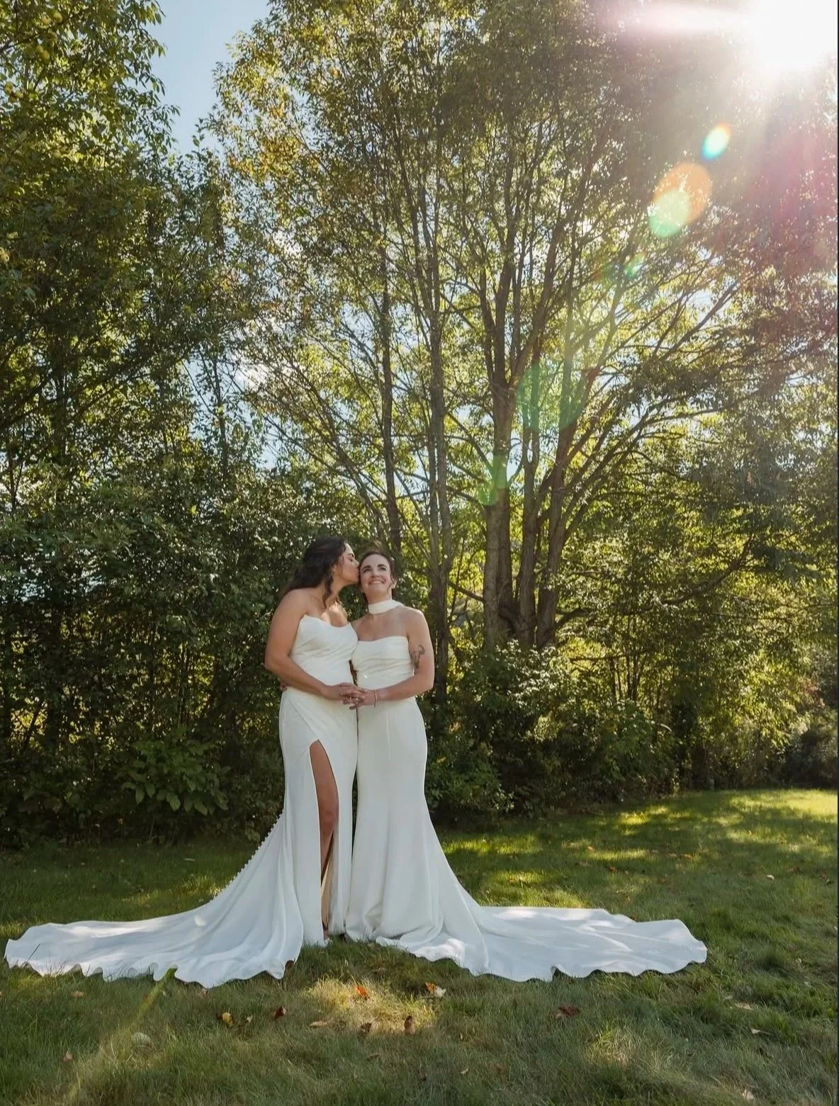 Two brides in white wedding gowns embrace and pose outdoors in a sunlit garden.