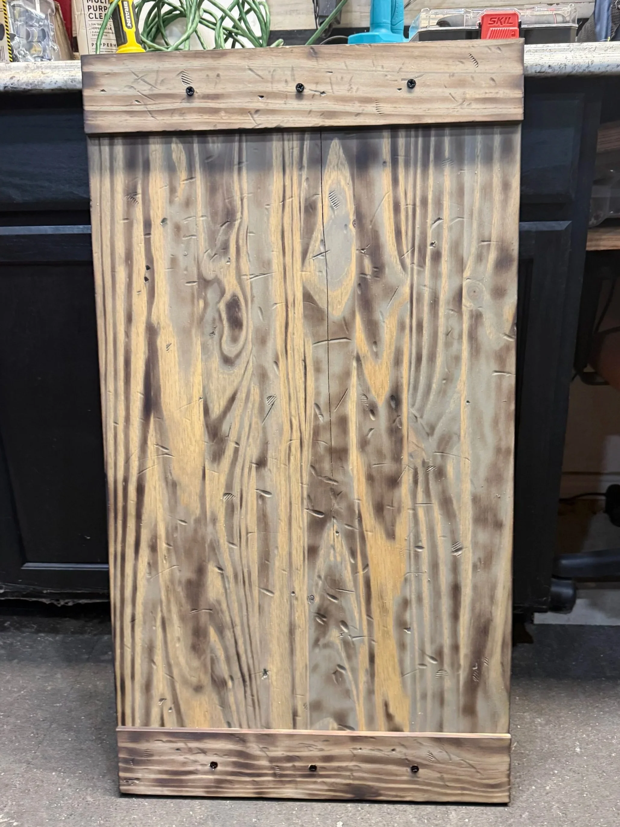 A wooden piece with a natural finish, featuring visible grain, knots, and carved patterns, leaning against a black surface. It has black screws at the top and bottom. The background includes various tools and supplies on a workbench.