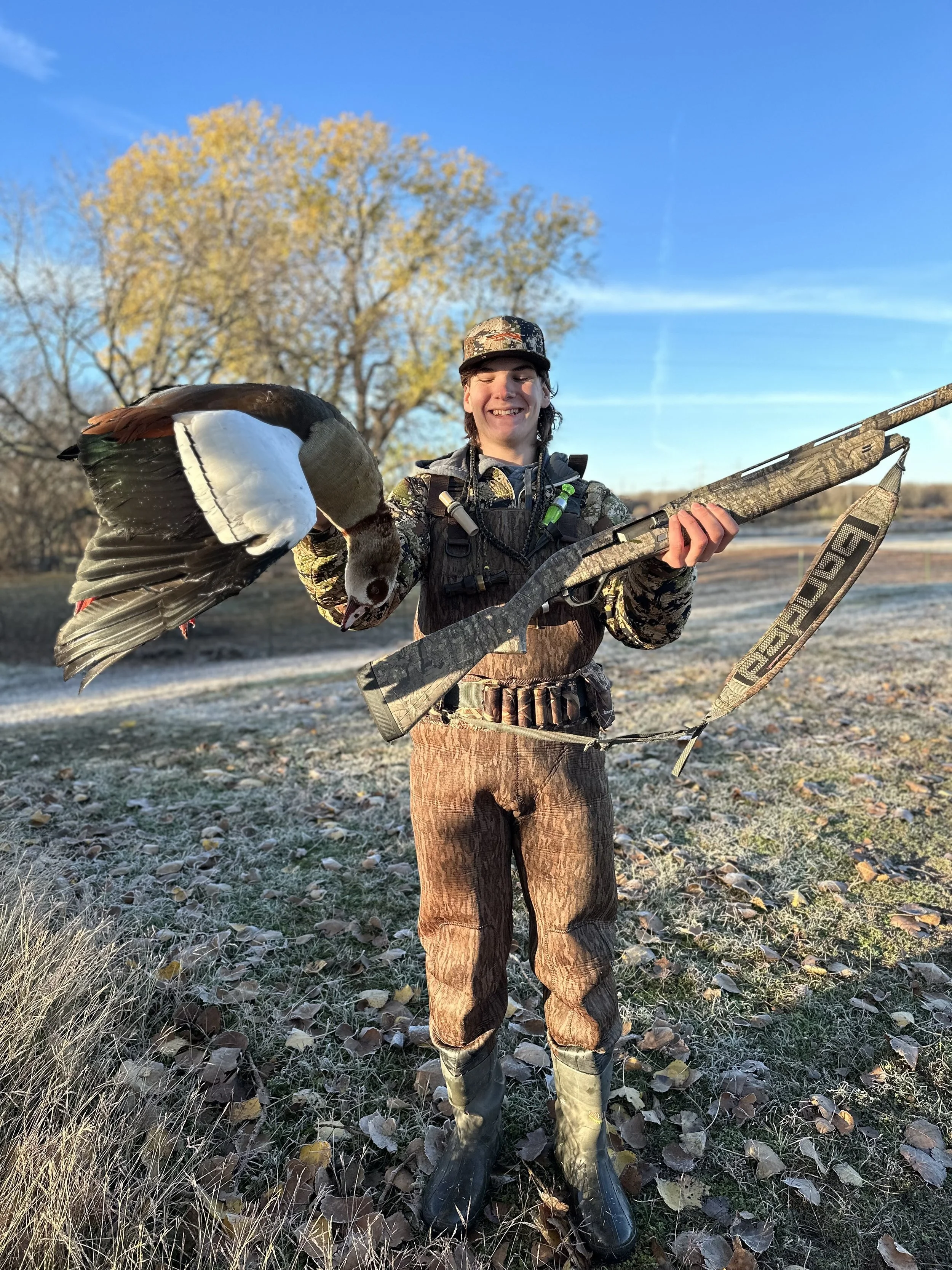 A person in camouflage clothing holding a duck and a hunting rifle outdoors during fall with trees and grass in the background.