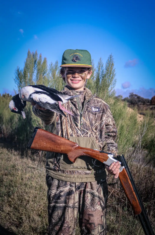 Young person dressed in camouflage hunting gear holding a duck and a shotgun outdoors with trees and a blue sky in the background.