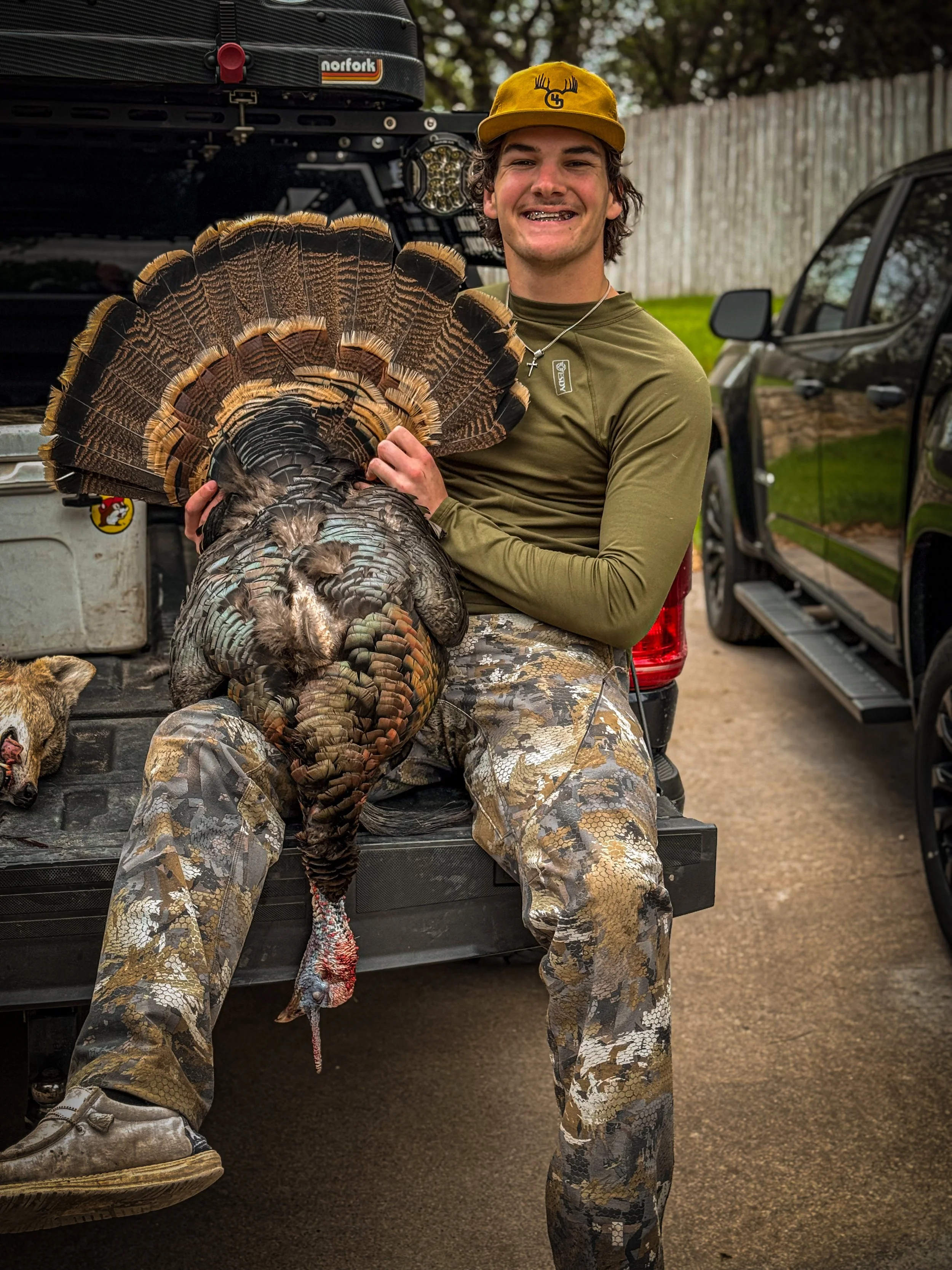 Young man sitting in the back of a truck holding a large wild turkey he hunted. The young man is smiling and wearing camouflage pants, a green long-sleeve shirt, a yellow cap, and a cross necklace. There are hunting-related equipment and vehicles visible in the background.