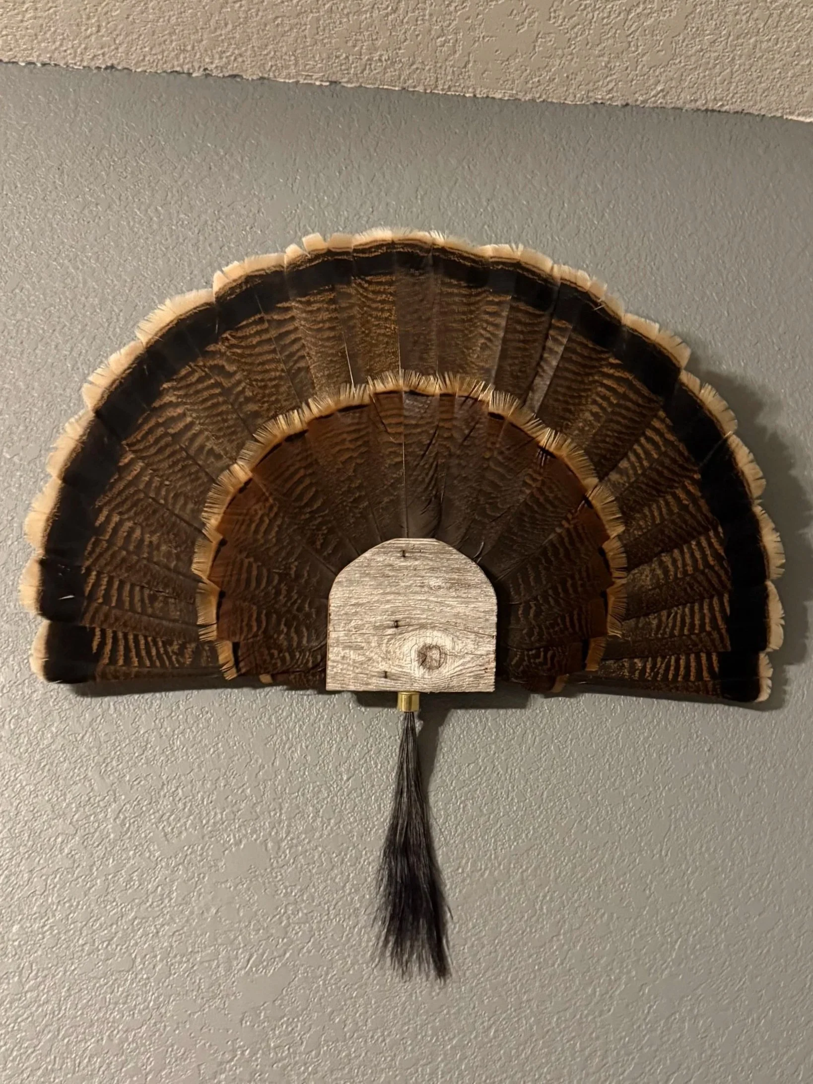 A decorative wall hanging in the shape of a fan with a wood grain design, black accents, fringes, and a tassel at the bottom, mounted on a gray textured wall.