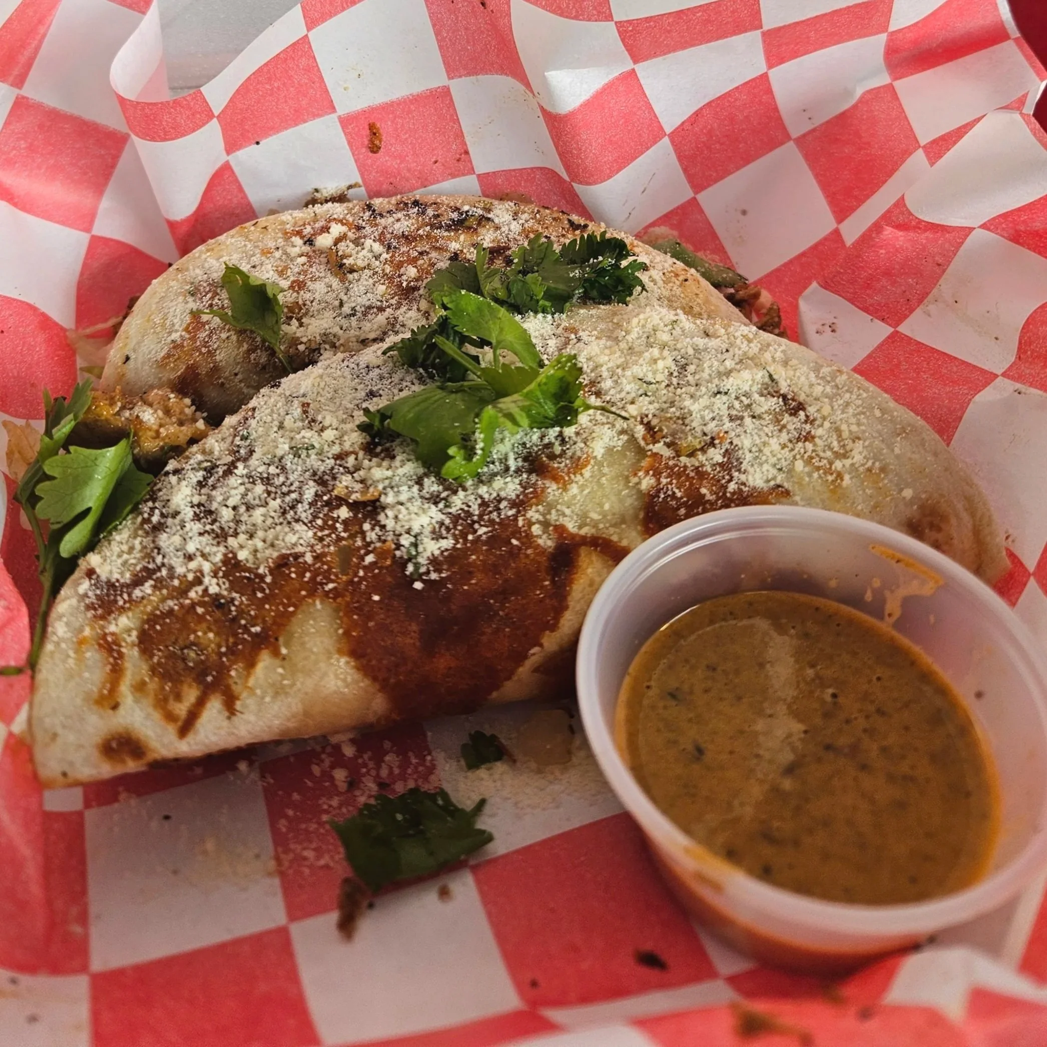 Close-up of a burrito wrapped in white paper with red checkered pattern, garnished with fresh cilantro, with two small containers of sauce on the side.