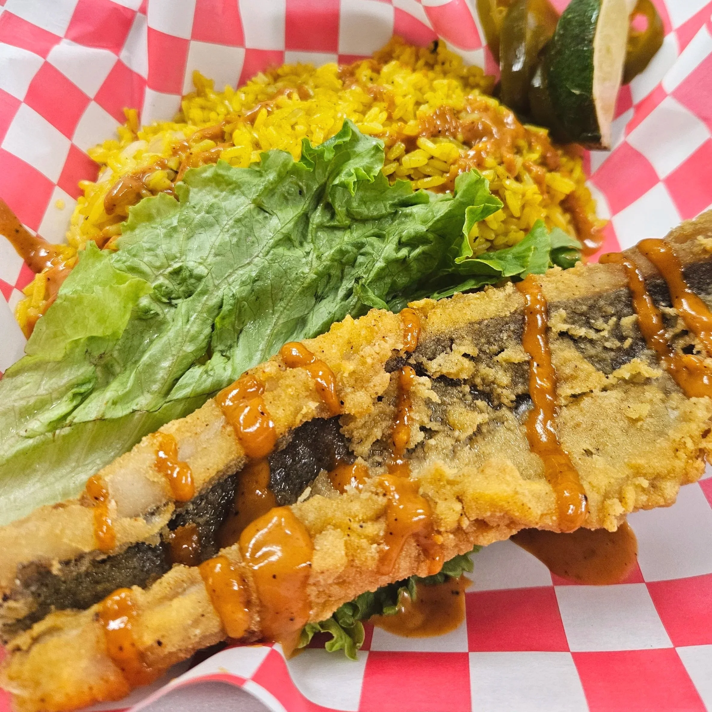 Close-up of fish fillet with breading and orange sauce, with leaf lettuce on side, in a red and white checkered food basket.