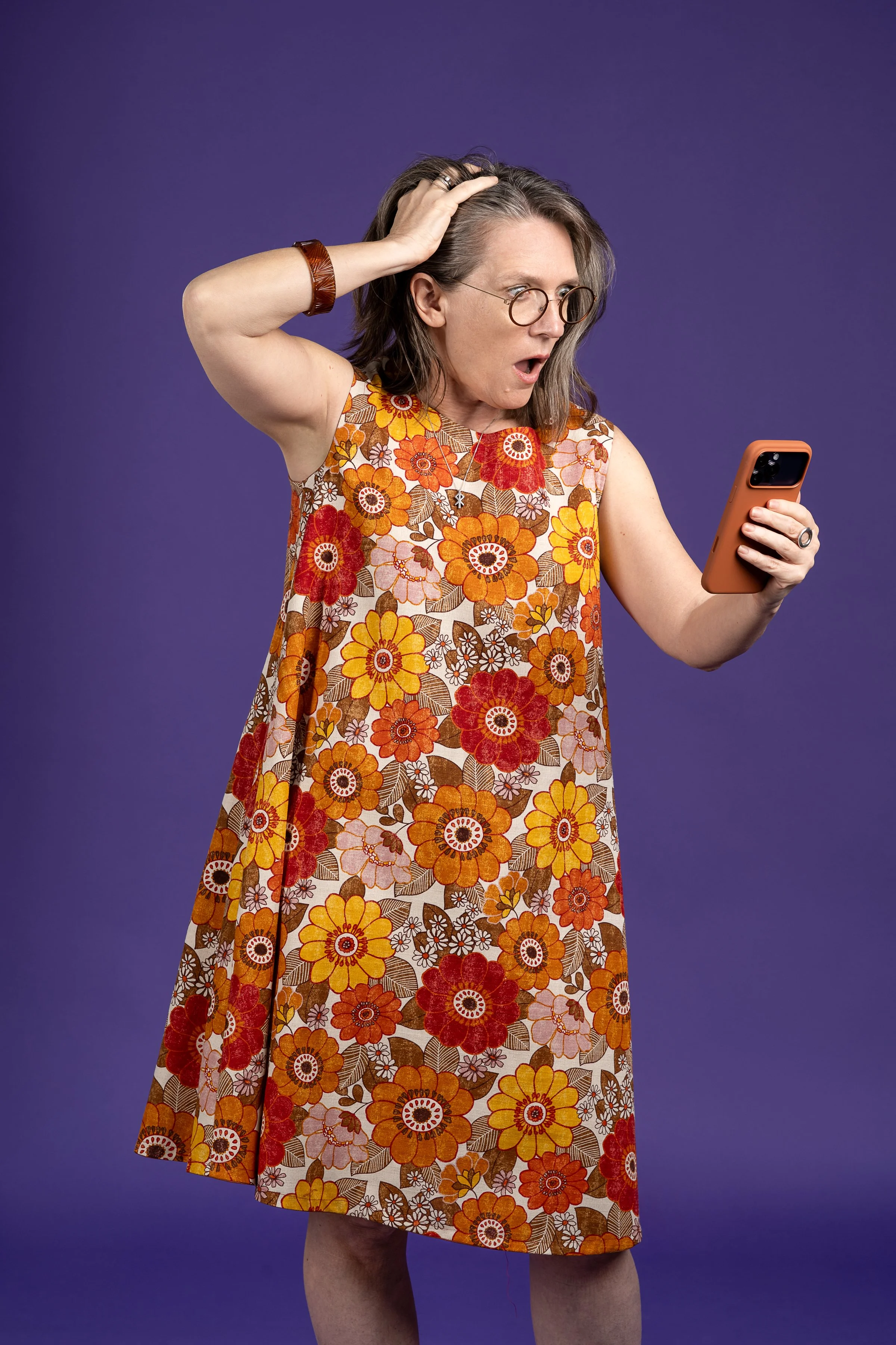 Surprised woman wearing glasses in a floral dress looking at a smartphone against a purple background.