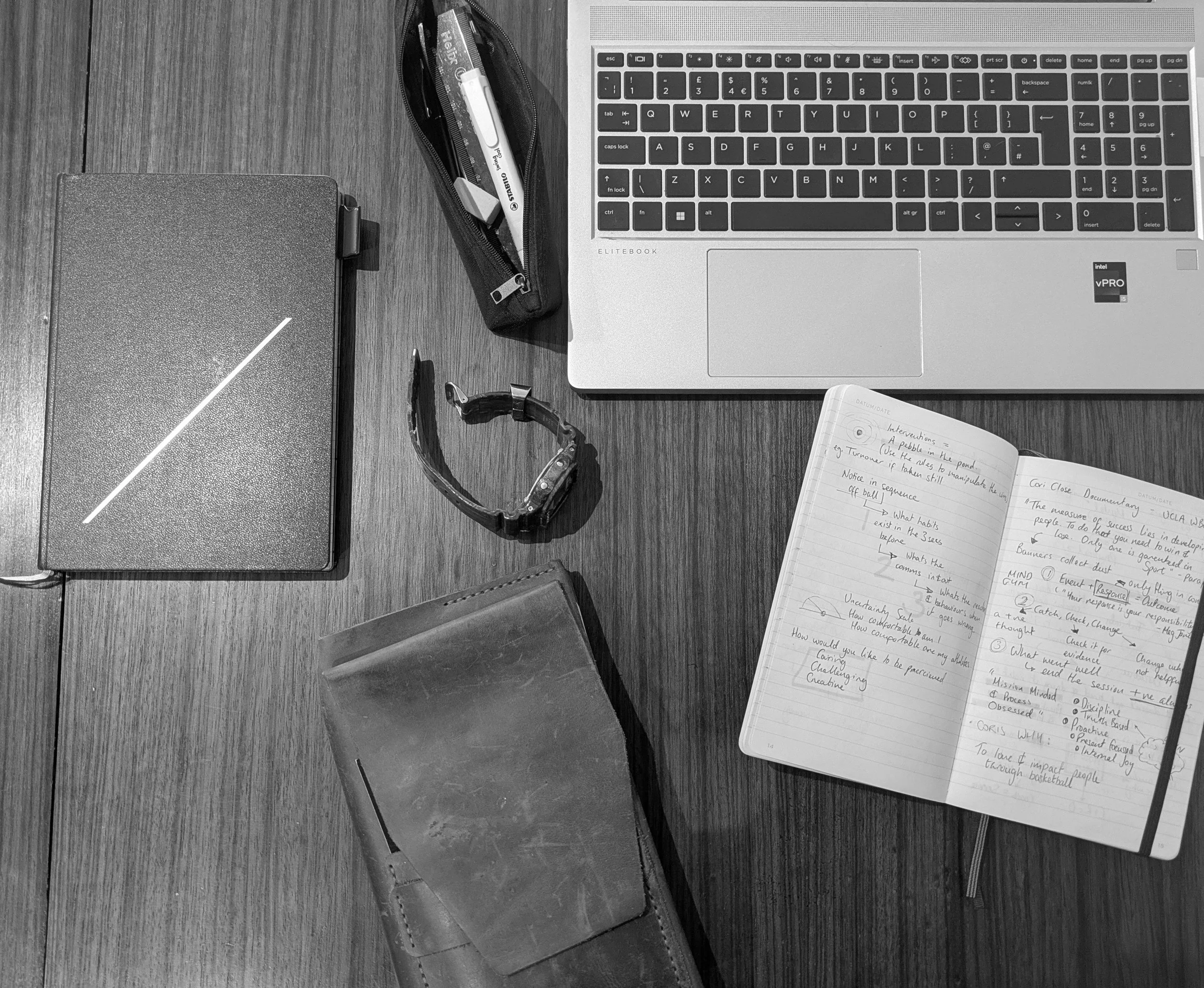 Black-and-white overhead view of a sports practitioner's desk, featuring a laptop and the physical four-notebook system used for reflective practice and mentoring.