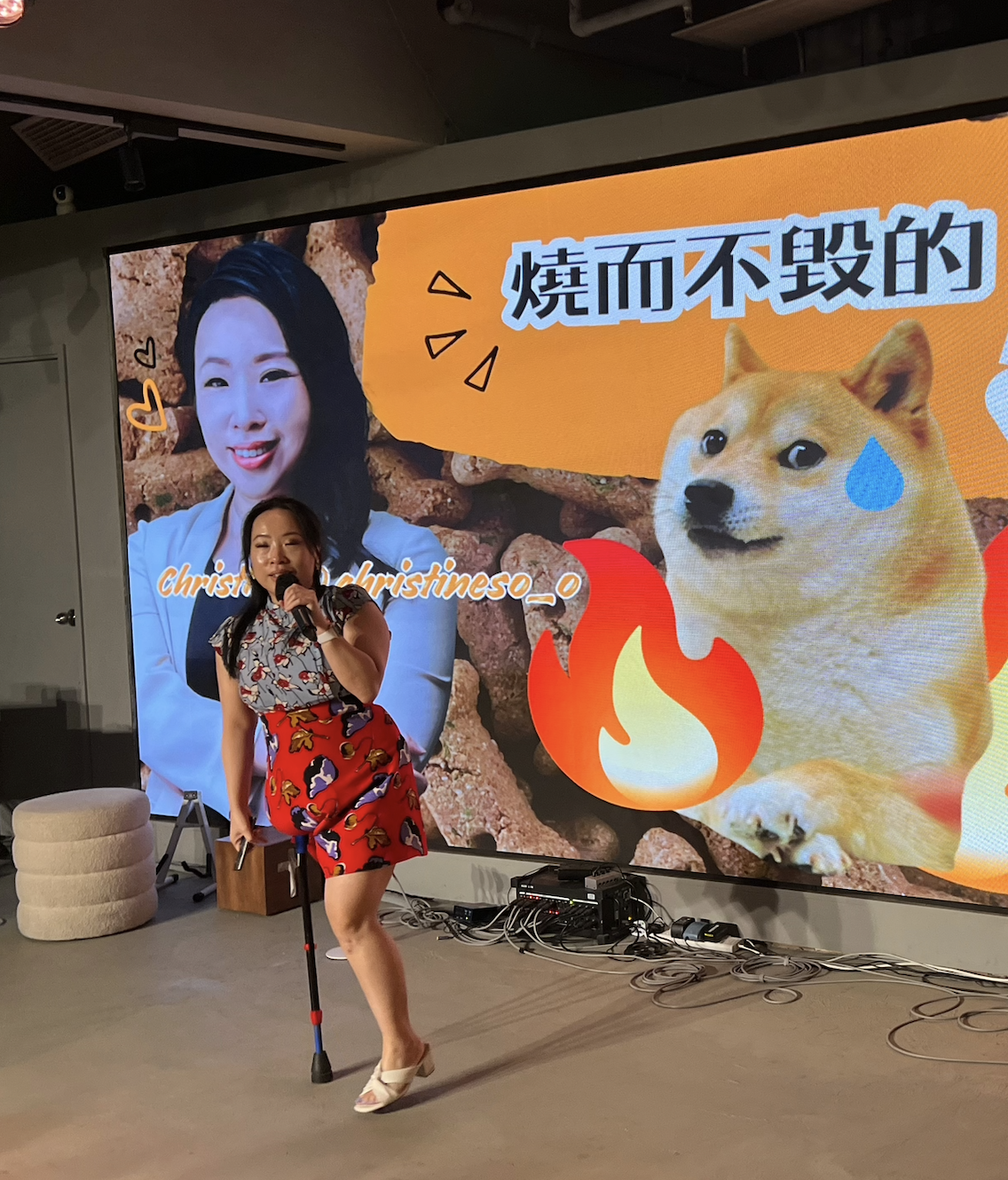 Woman speaking into a microphone in front of a large presentation screen showing a Shiba Inu dog meme, Chinese text, and a social media handle.