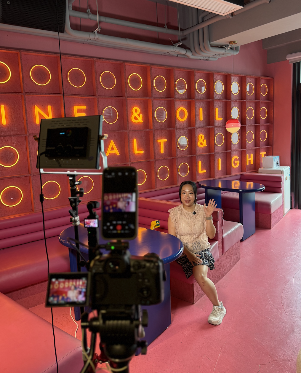 Woman waving at a camera while seated in a colorful pink lounge with neon wall lettering and filming equipment in the foreground