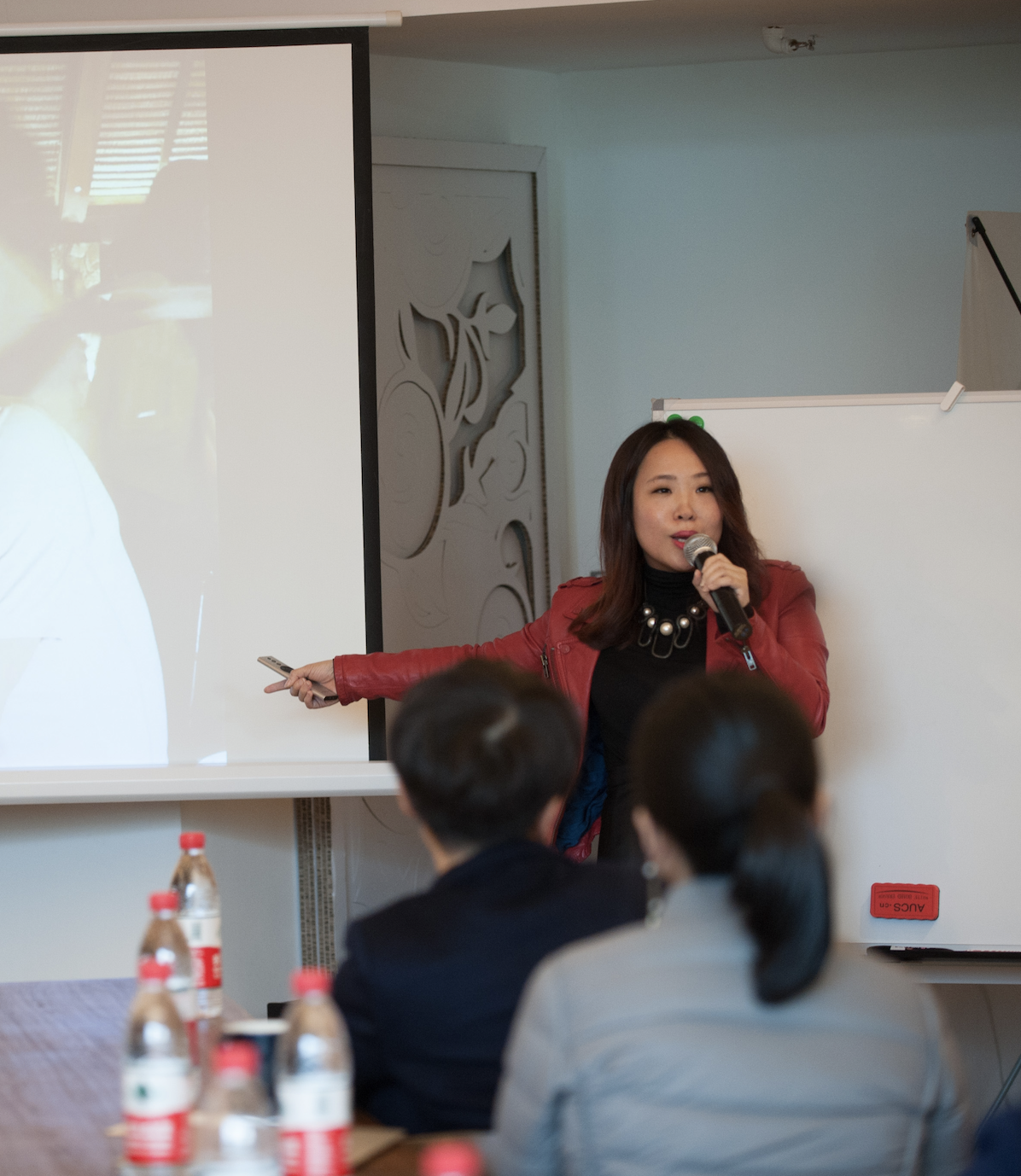 Woman in a red jacket speaking into a microphone during a presentation to an audience