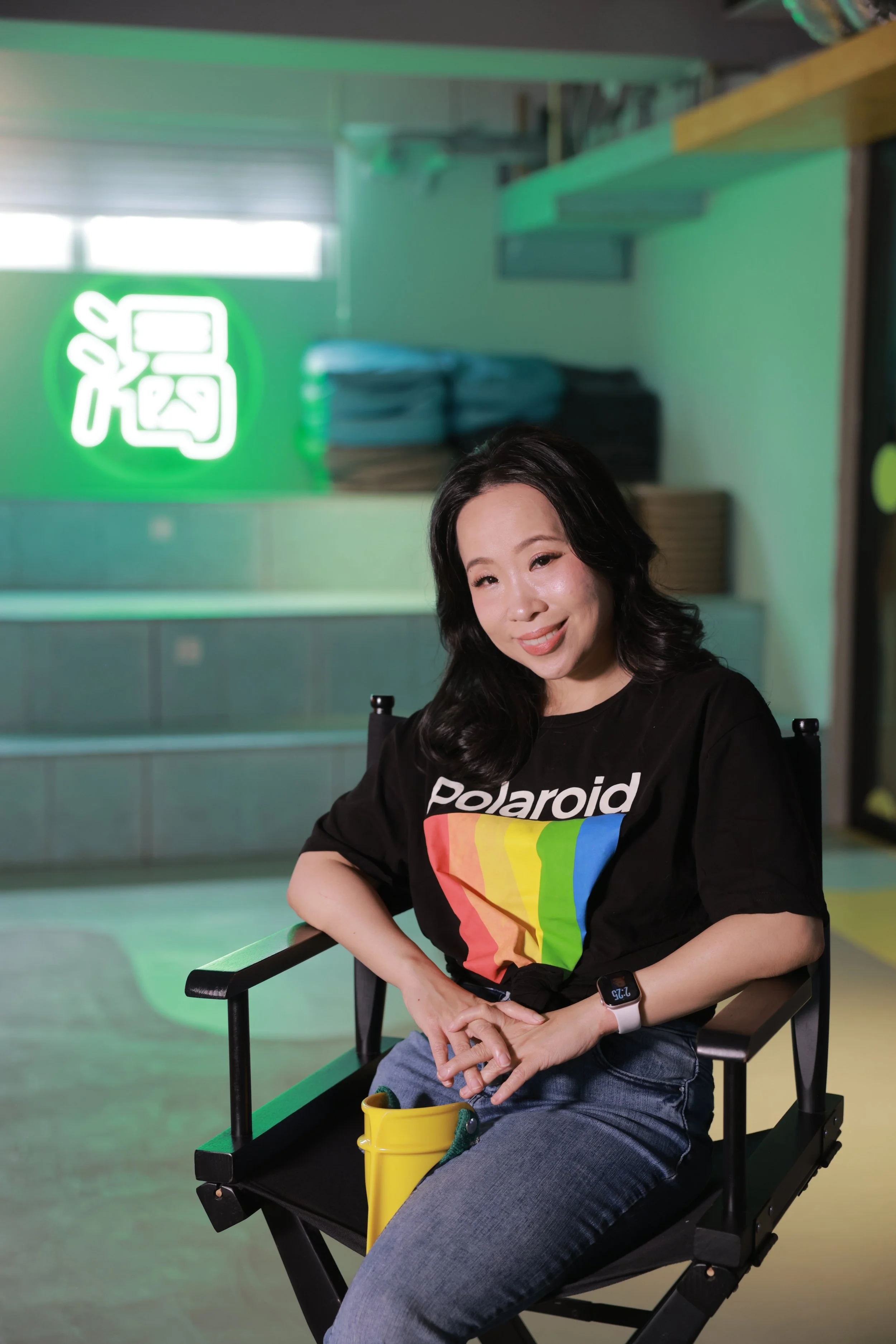 Smiling woman in a black Polaroid T-shirt seated in a director’s chair beside a yellow mug.