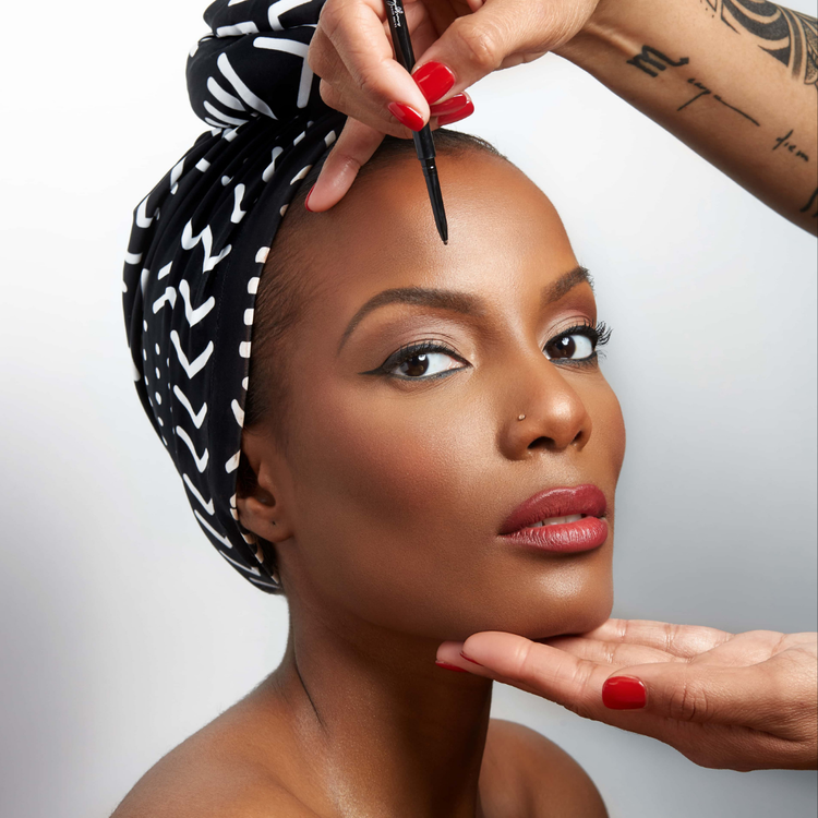 A woman with a headwrap getting her eyebrows and makeup done by a makeup artist