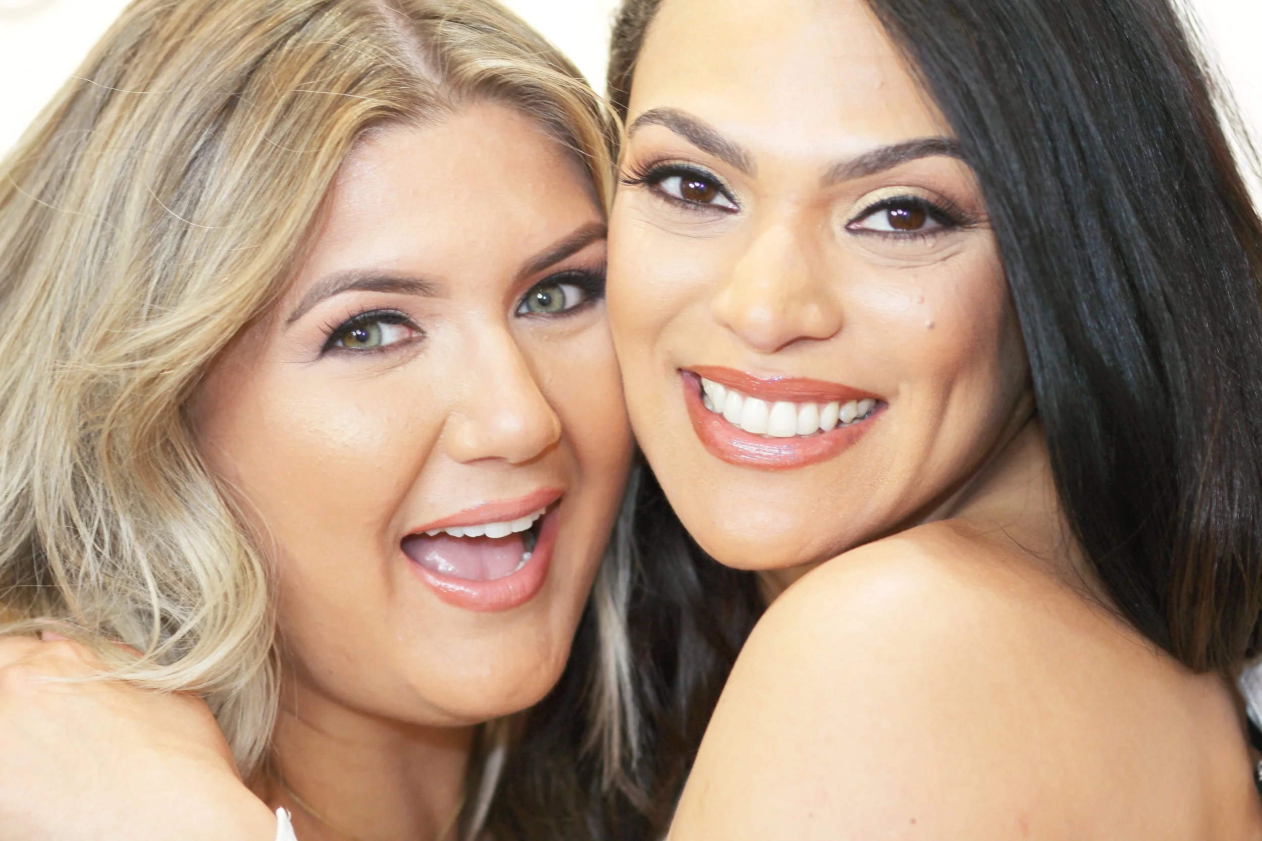 Two smiling women with close-up faces, one with blonde hair and light eyes, the other with dark hair and brown eyes.