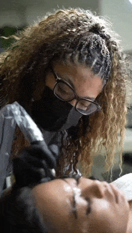 A woman with curly hair and glasses is performing a cosmetic tattooing procedure on a woman's eyebrows in a clinic setting.