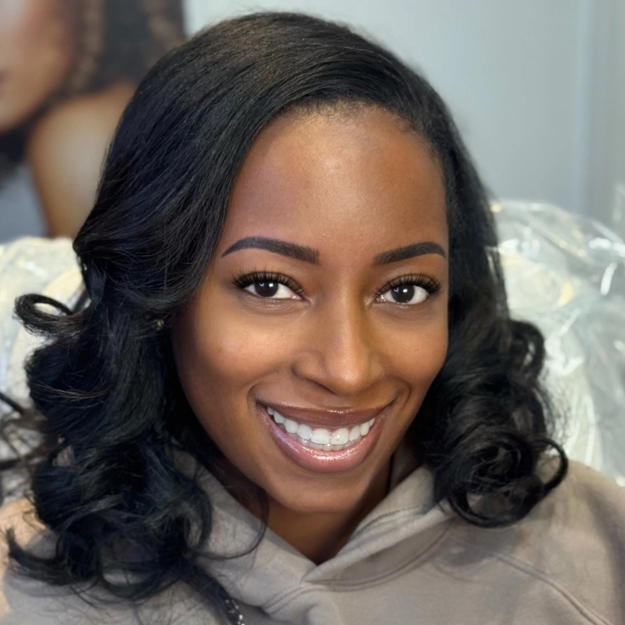Close-up of a smiling woman with wavy black hair, wearing makeup including mascara, eyeliner, and lipstick, in an indoor setting.