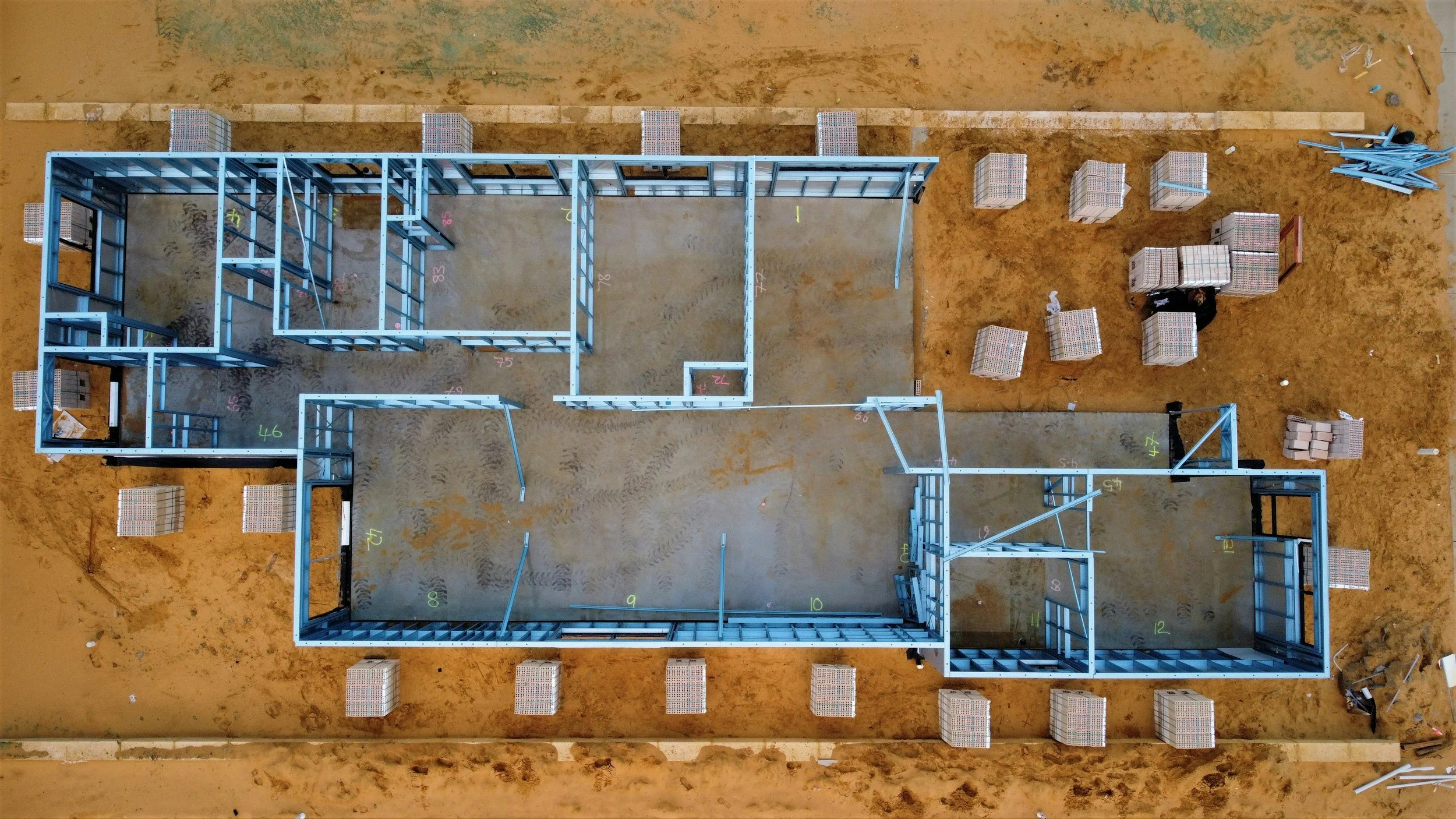 Aerial view of a steel-framed house under construction on a sandy construction site, with pallets of bricks and building materials nearby.