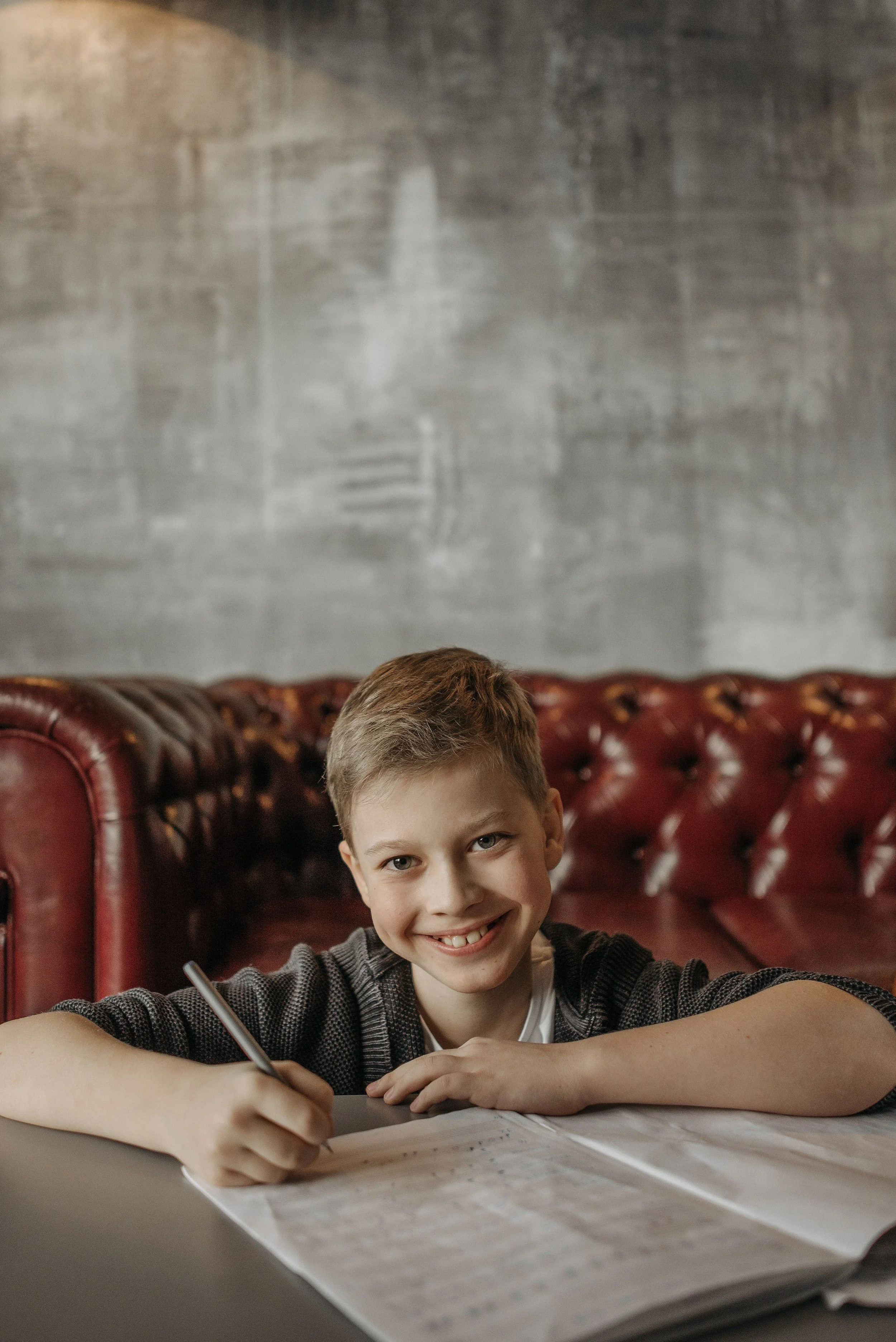 A young boy with short light brown hair and blue eyes sitting at a table, smiling, holding a pen, with a large booklet or worksheet in front of him. He is wearing a striped gray and black sweater. Behind him is a red leather tufted booth and a textured gray wall.