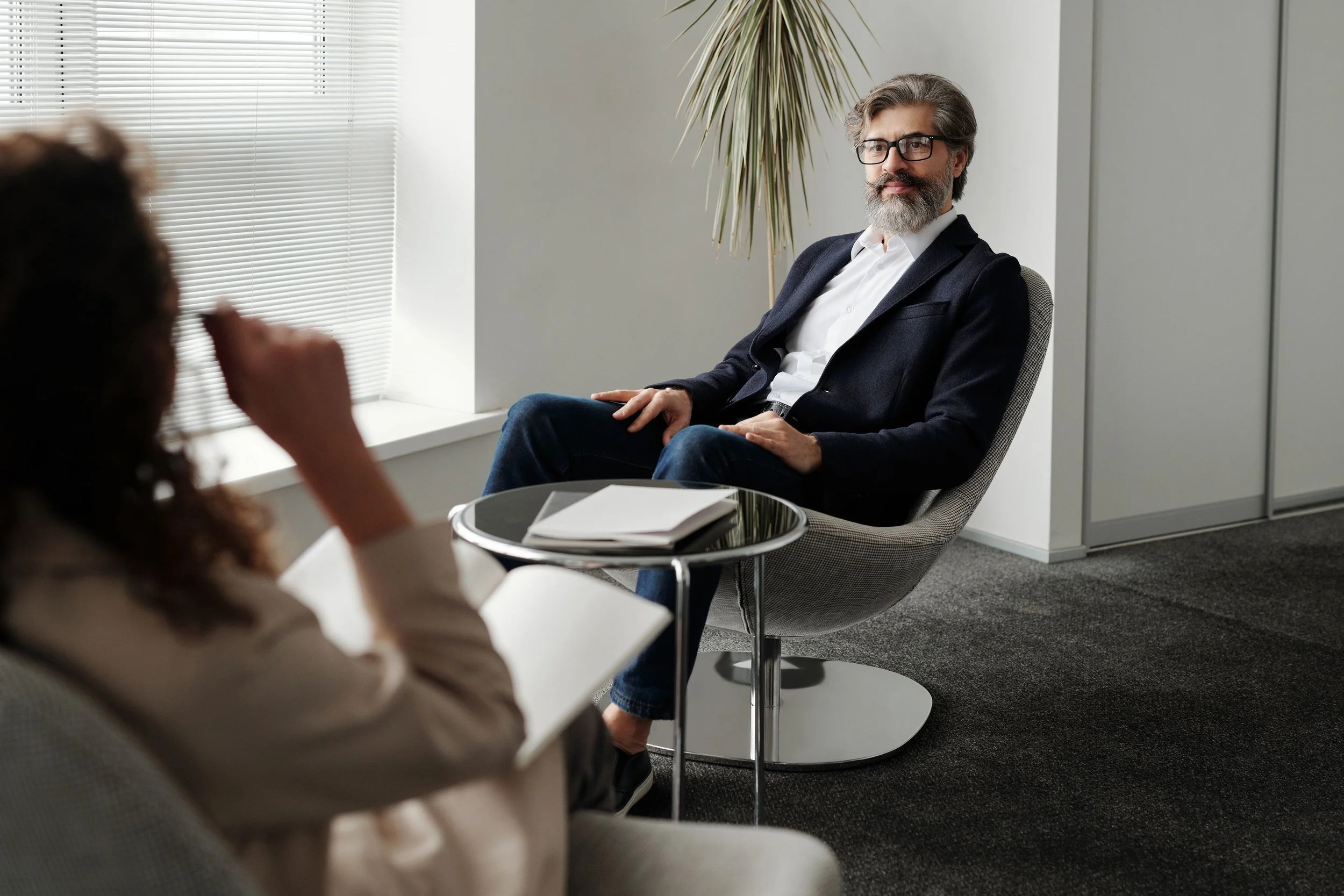 A man with gray hair, beard, and glasses sitting in an office chair, facing another person who is out of focus. The man is wearing a dark blazer and white shirt, in a modern office with a potted plant, window with blinds, and a small round table with notebooks.