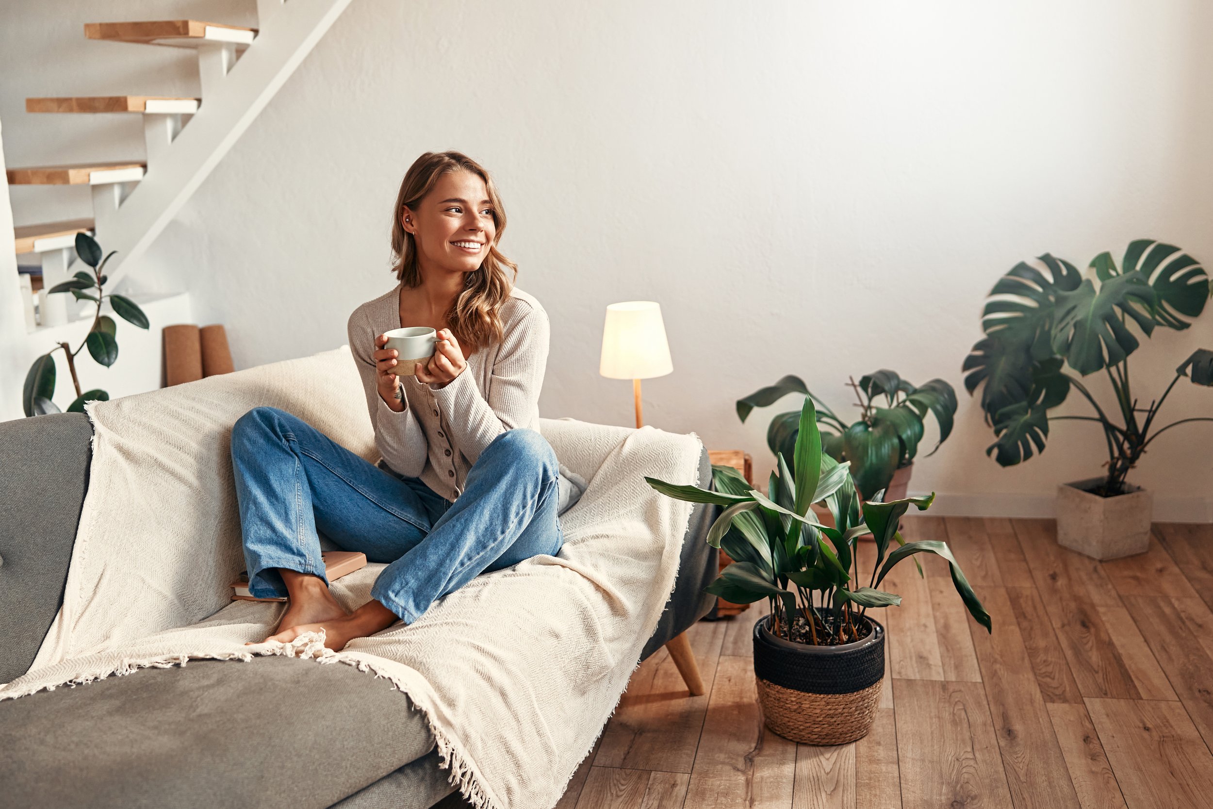 A young woman with light brown hair, wearing a beige cardigan and blue jeans, sitting on a beige sofa in a living room, holding a white mug, smiling and looking to the right among green houseplants.