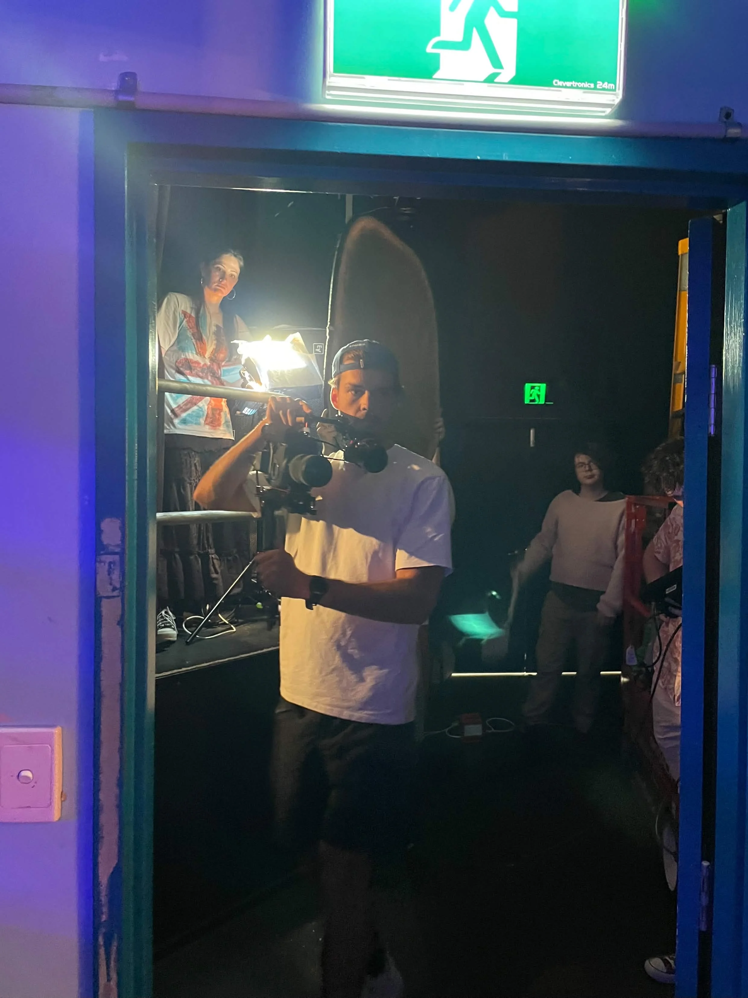 People gathered inside a dimly lit venue, with a man filming using a camera rig beneath a green emergency exit sign.