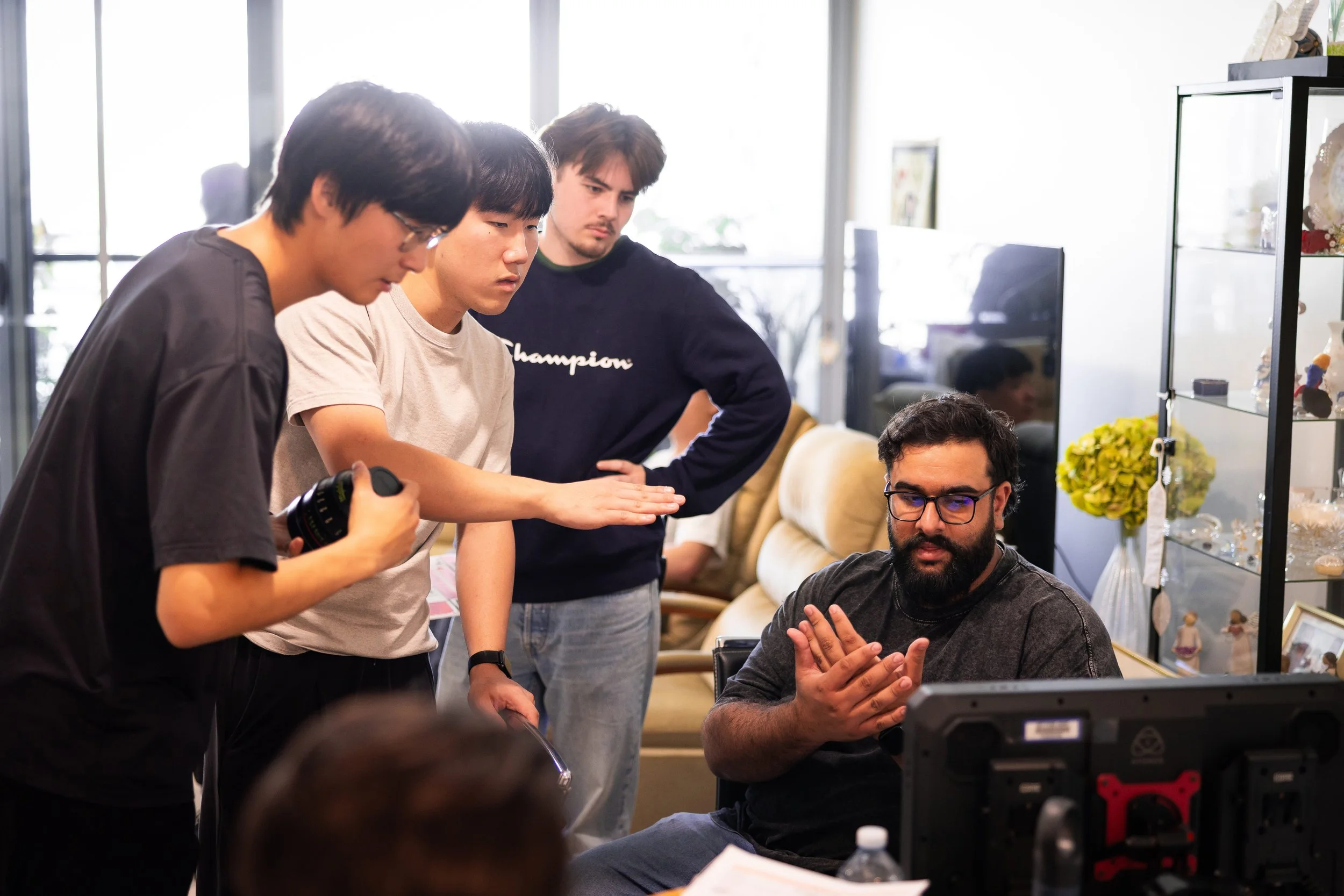 Film crew gathered around a seated man reviewing a monitor in a bright office or studio.