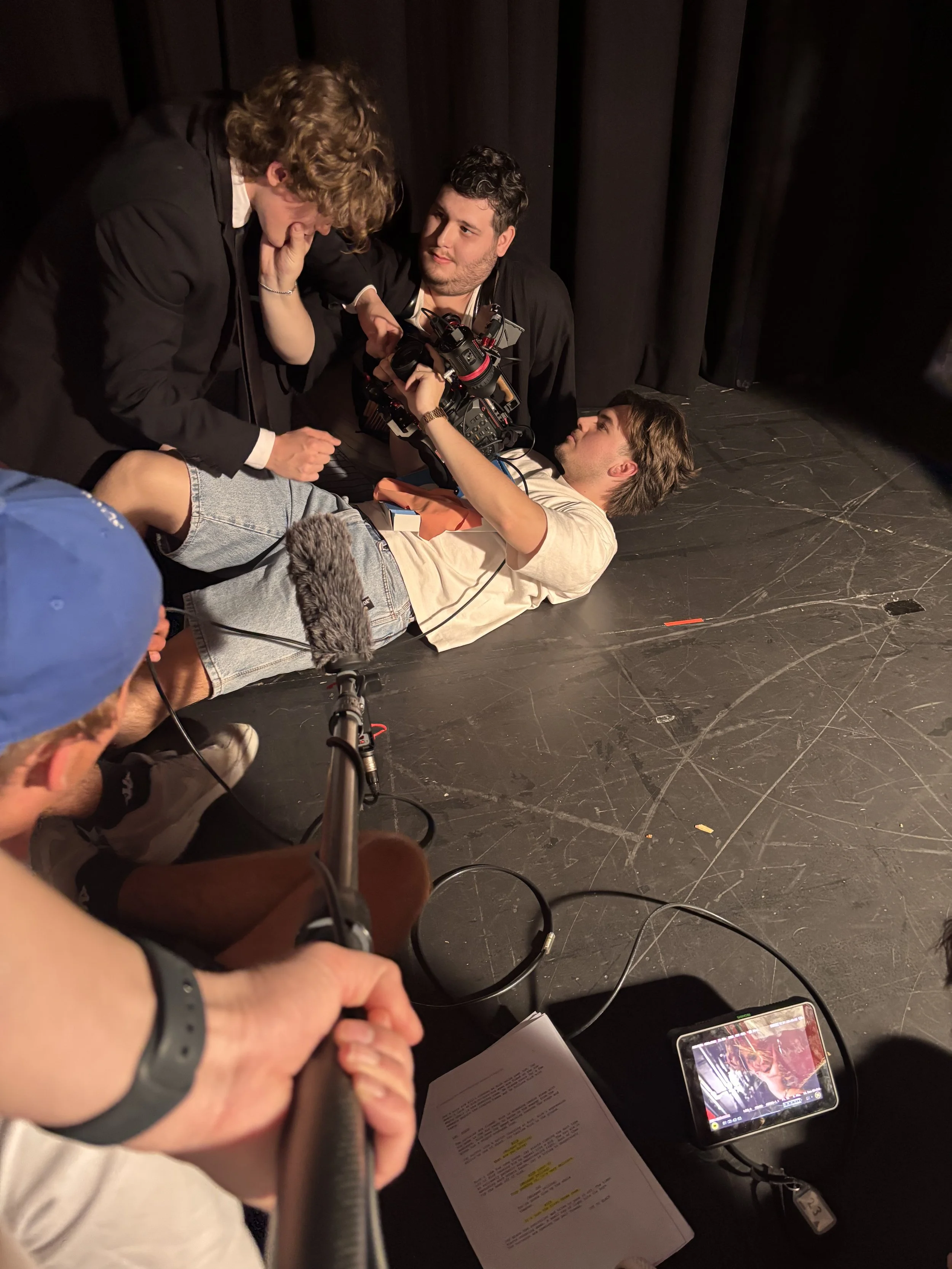 Film crew members operate a professional video camera on a theater stage behind black curtains.