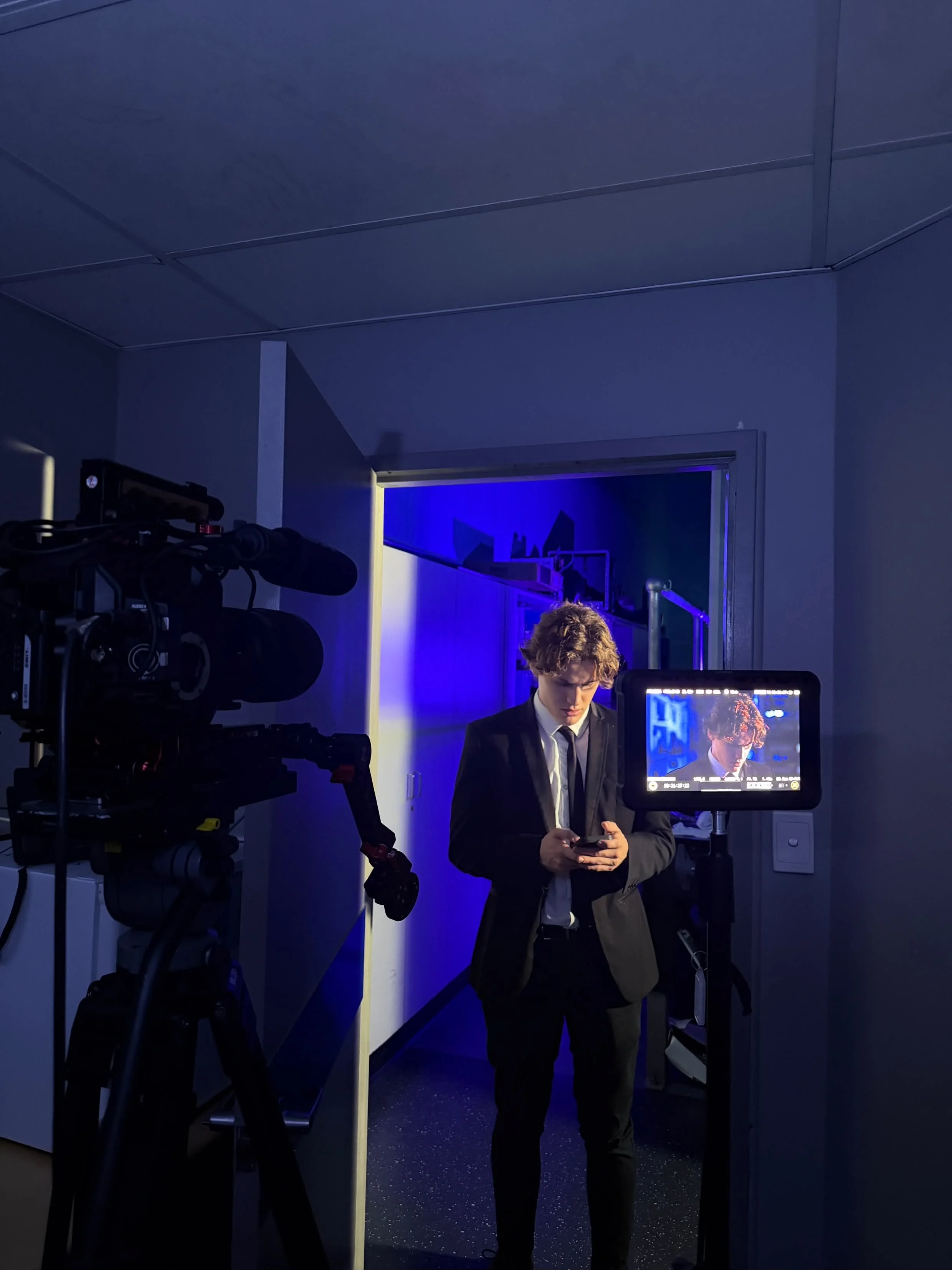 Film set with a young man in a suit checking his phone, a professional camera, and a monitor displaying his close-up.