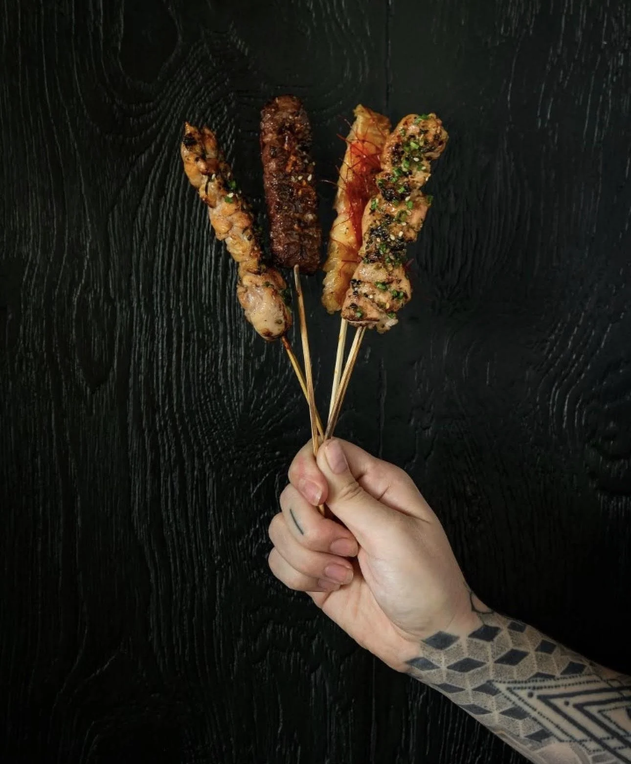 A hand with a tattoo holds four skewers of grilled meat against a black textured background.
