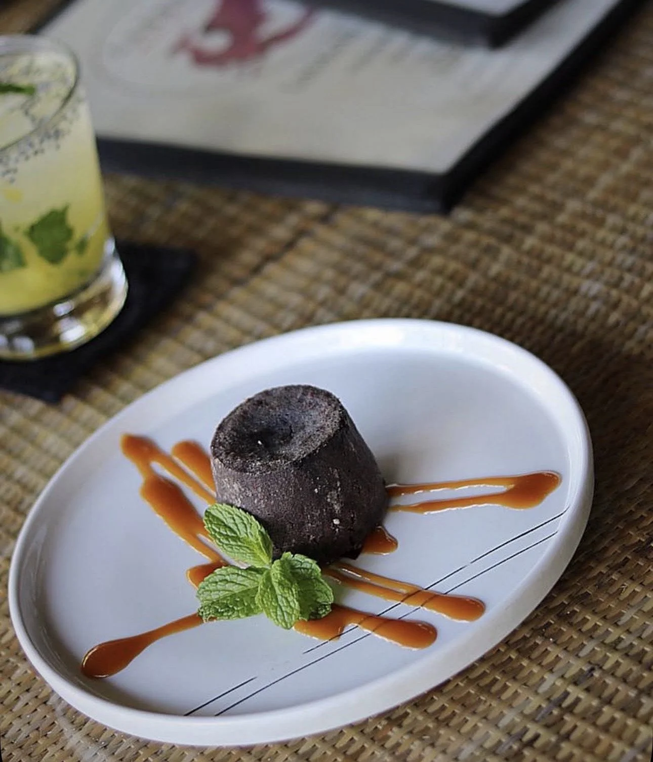 Chocolate dessert served on a white oval plate, garnished with caramel sauce and a sprig of mint, on a woven placemat.