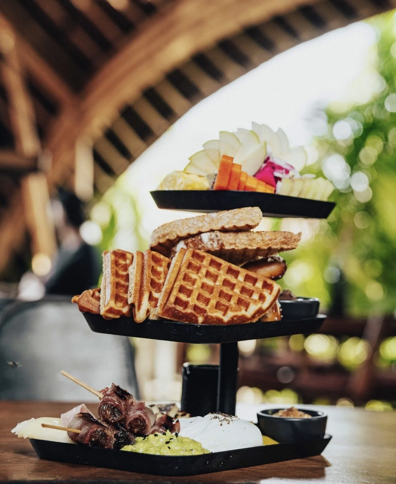 A three-tier black serving platter with waffle sandwiches, grilled meat, vegetables, and garnishes, set on a wooden table outdoors with a blurred background of trees and a wooden roof.