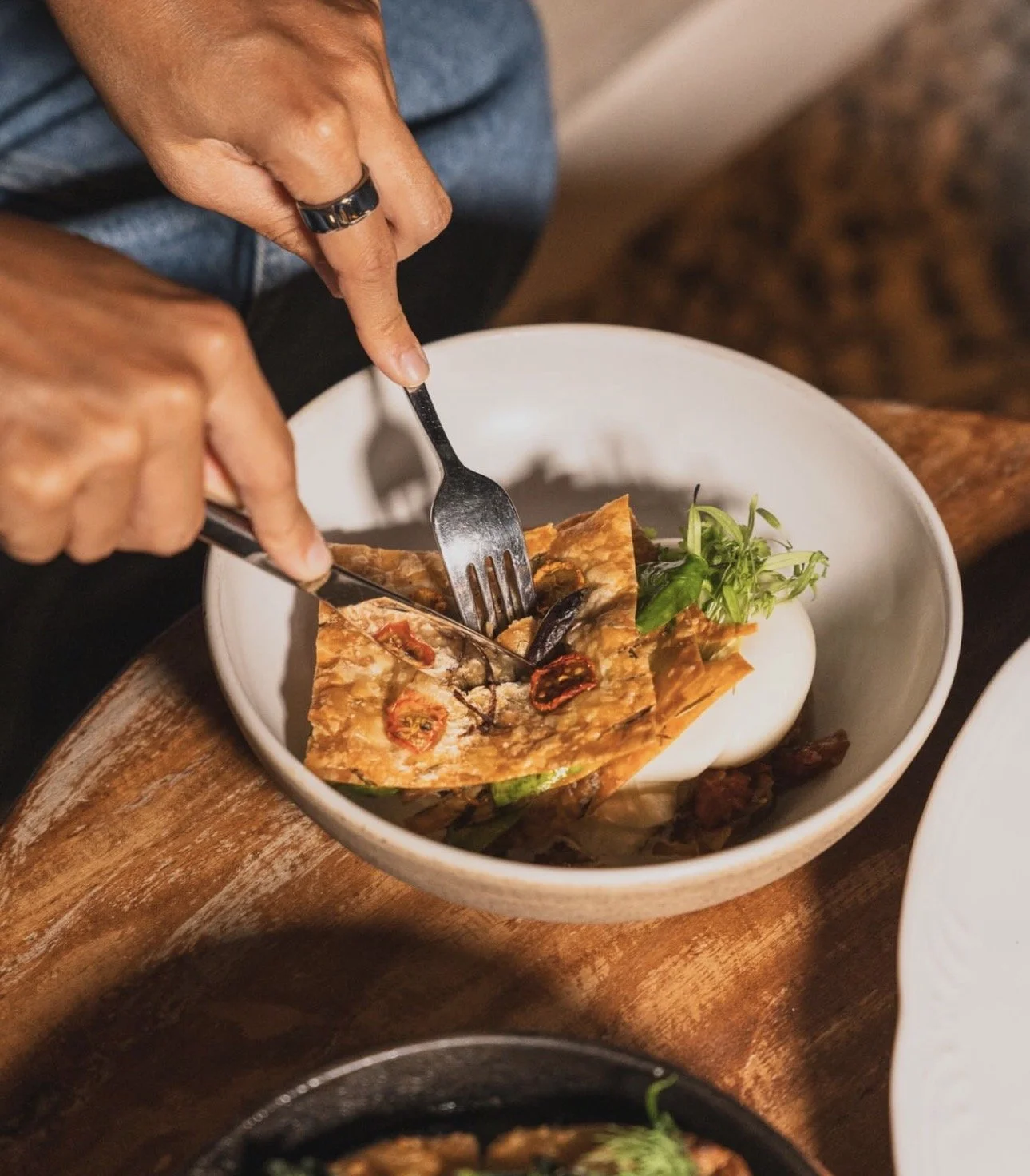 Person using fork and knife to eat a slice of pizza with sun-dried tomatoes and greens in a white bowl.