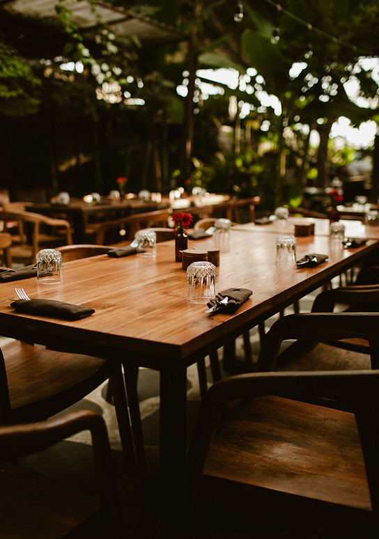 Empty dining table set with glasses, black napkins, utensils, and a small red flower centerpiece in an outdoor restaurant or patio area.