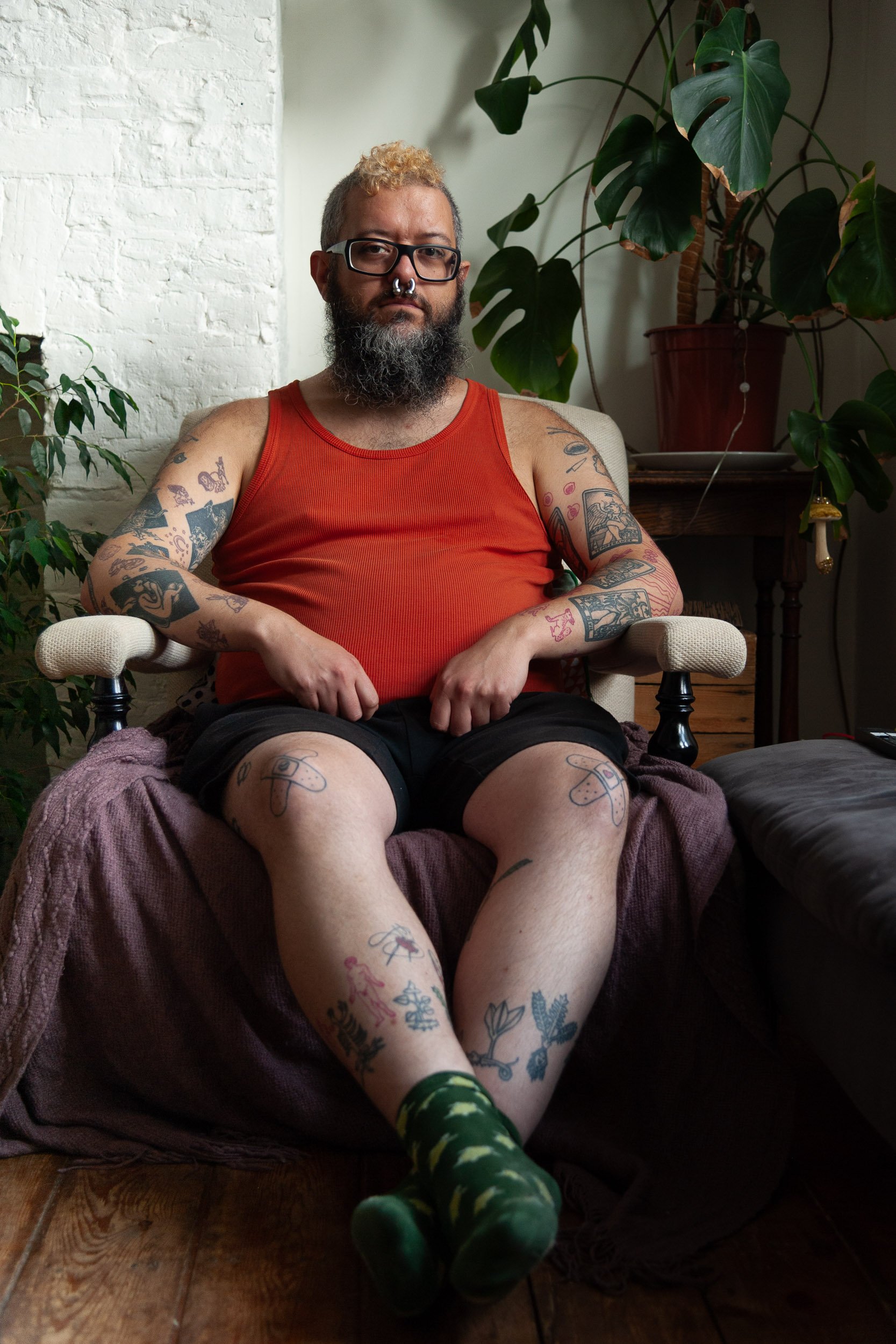 Tattooed bearded man wearing glasses and a red tank top sits in an armchair surrounded by houseplants.