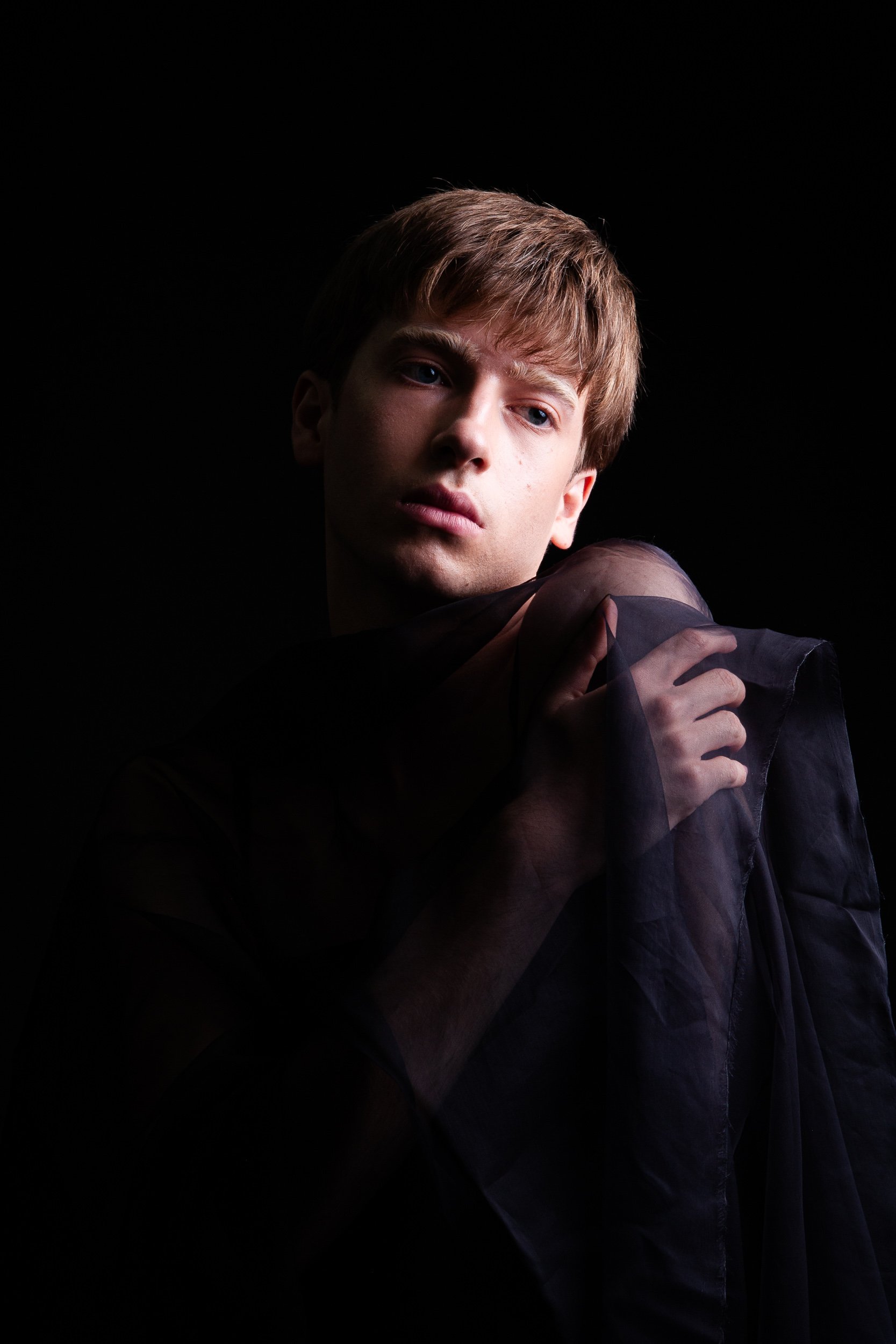 Portrait of a young man with tousled brown hair and a serious expression against a black background