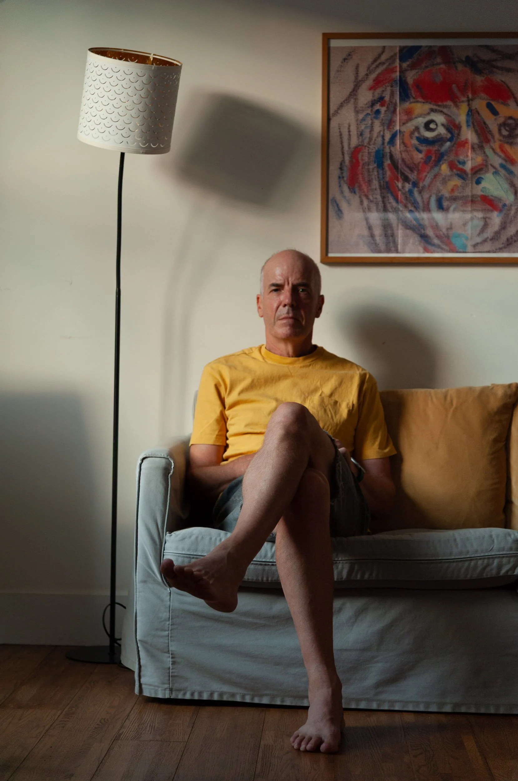 Man in a yellow shirt sitting barefoot on a couch beside a floor lamp and colorful framed artwork