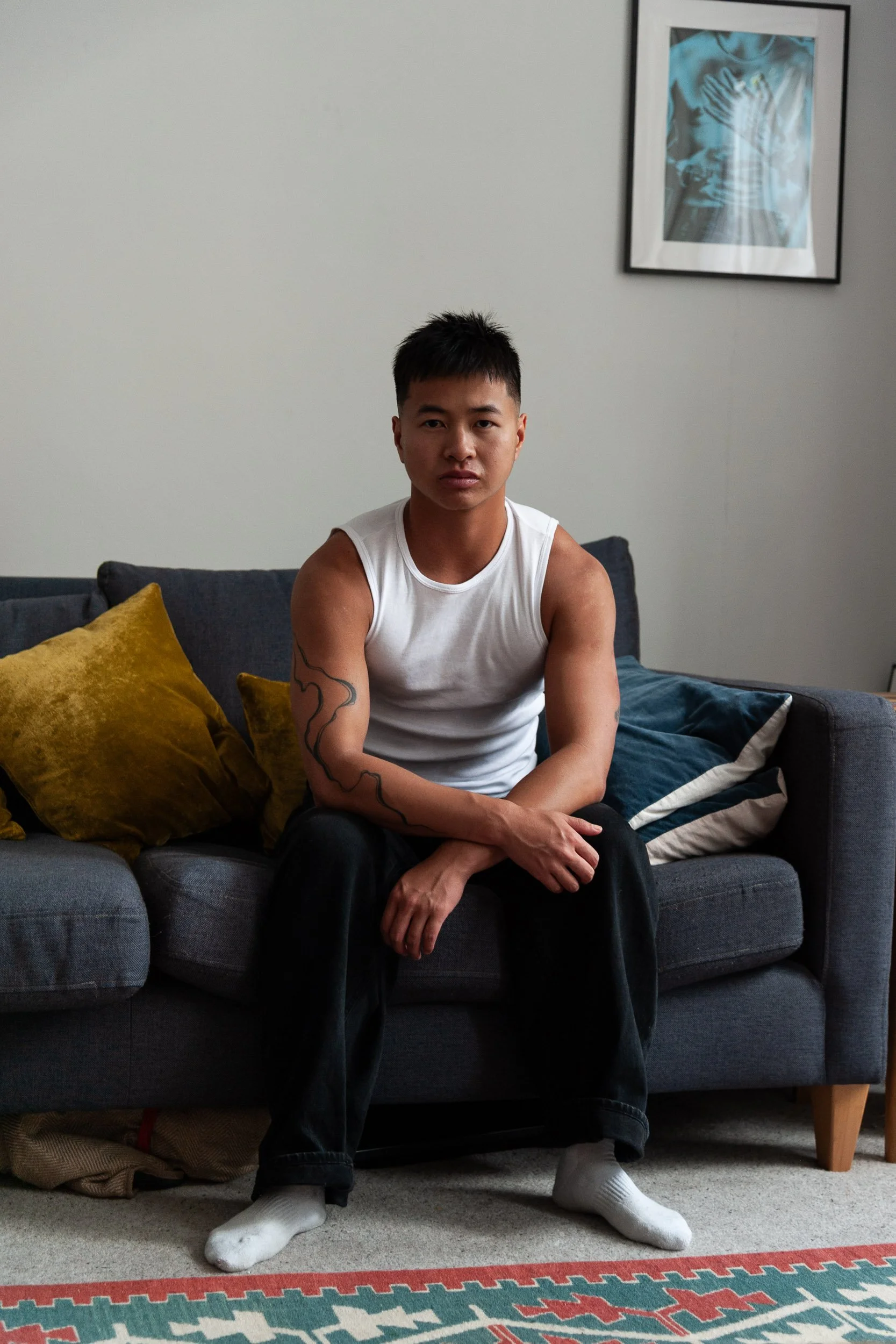 Young man with a tattoo sitting on a gray sofa in a living room, wearing a white tank top, black pants, and white socks