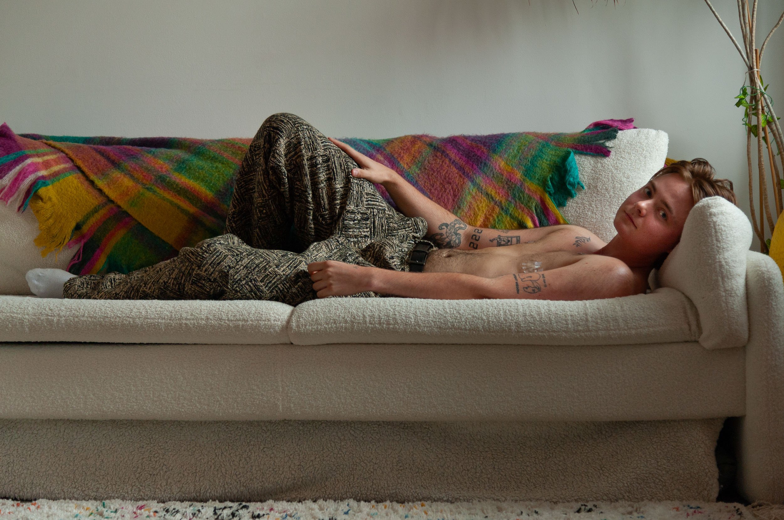 Tattooed shirtless person reclining on a white couch beneath a colorful plaid blanket, with a stuffed teddy bear beside them.