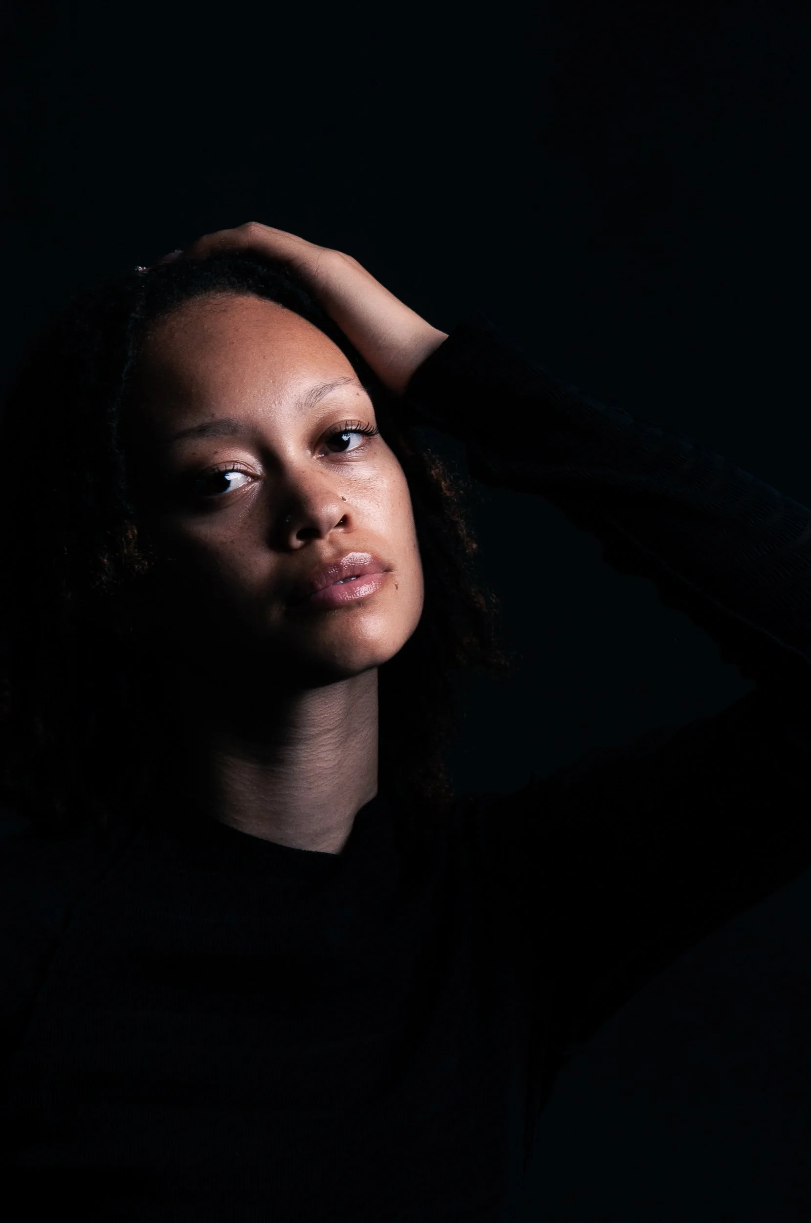 Close-up portrait of a Black woman with curly hair resting her hand on her head against a dark background